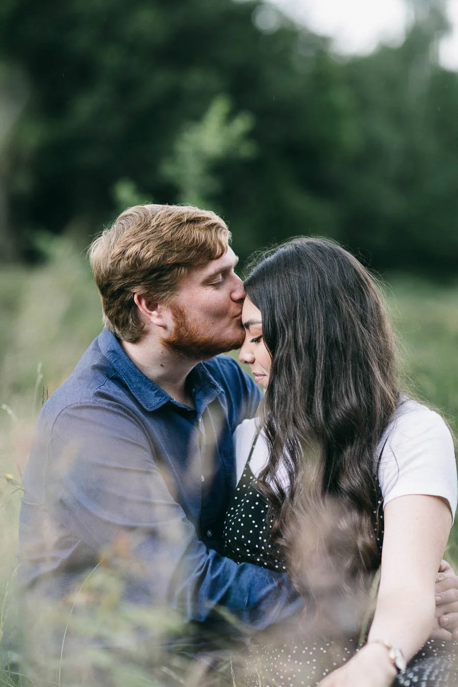 Engaged couple sitting in a field, man kissing his fiancée on the forehead. Tall grass blurred in the foreground. 