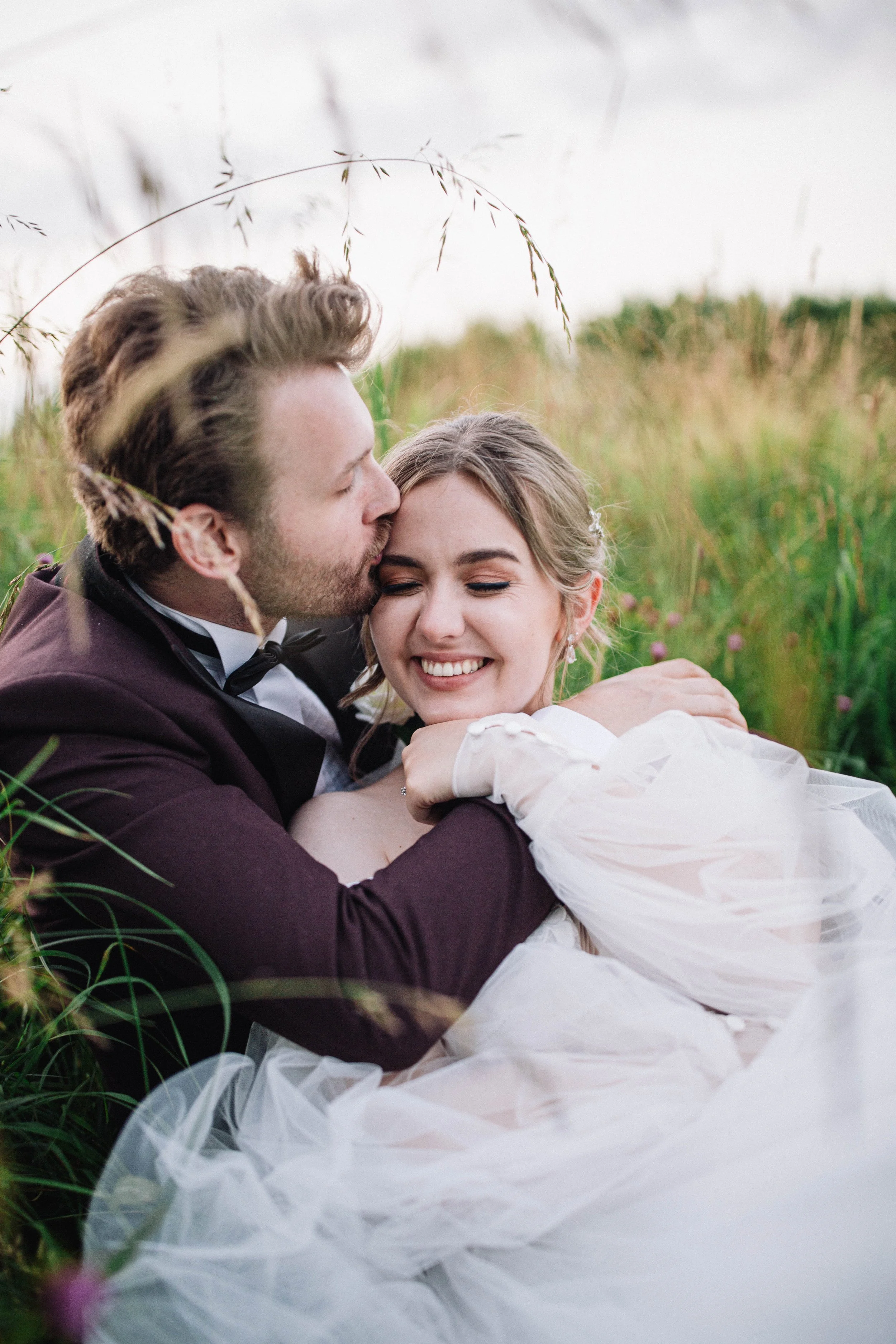 Bride and groom sitting in a grassy field. Groom kisses bride on the side of the head as she smiles.