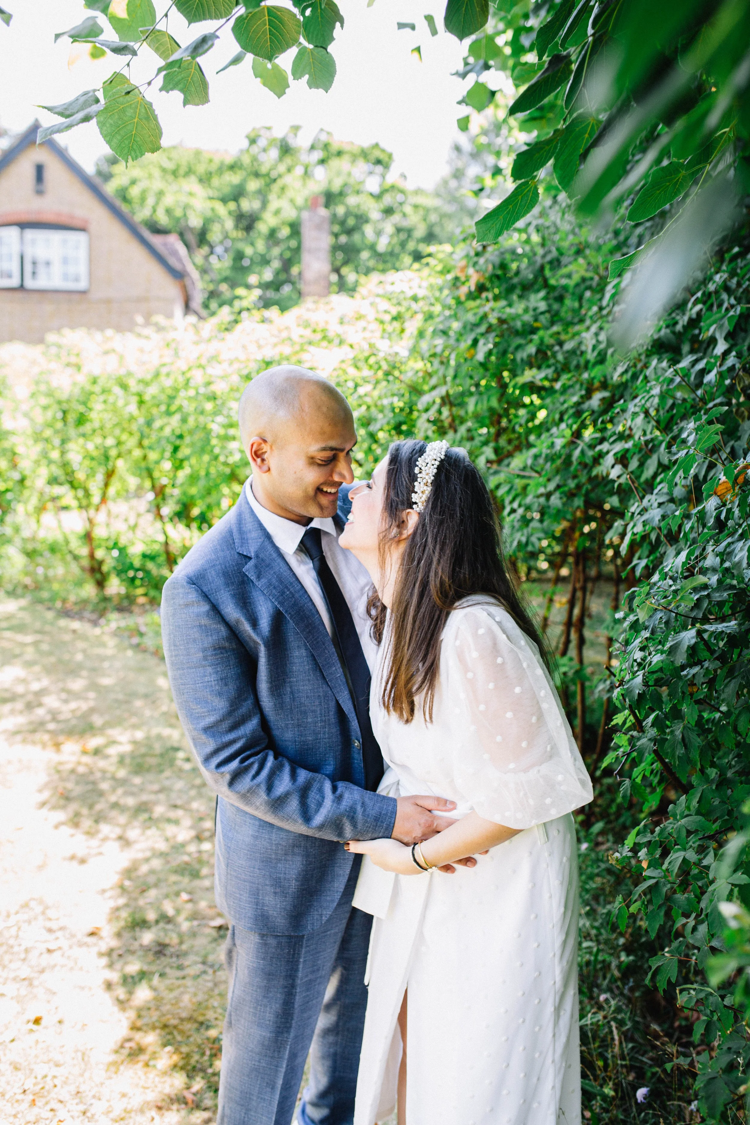 Newlywed couple embracing and smiling on a leafy garden path, with the bride wearing a white dress and floral hair accessory.