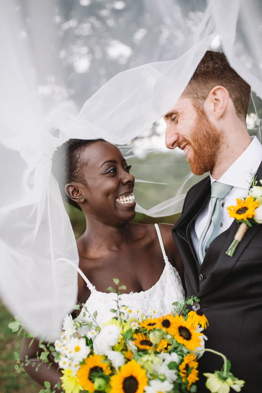 Bride and groom smile at each other with the brides veil draped over them both.