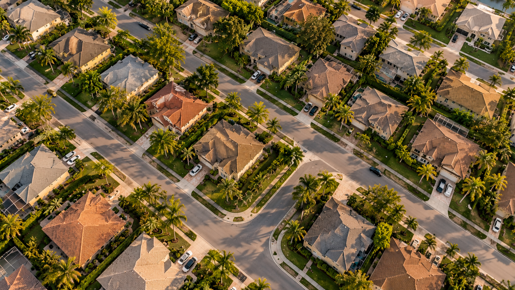 Aerial view of a suburban neighborhood with single-family homes, tree-lined streets, driveways, and parked cars.
