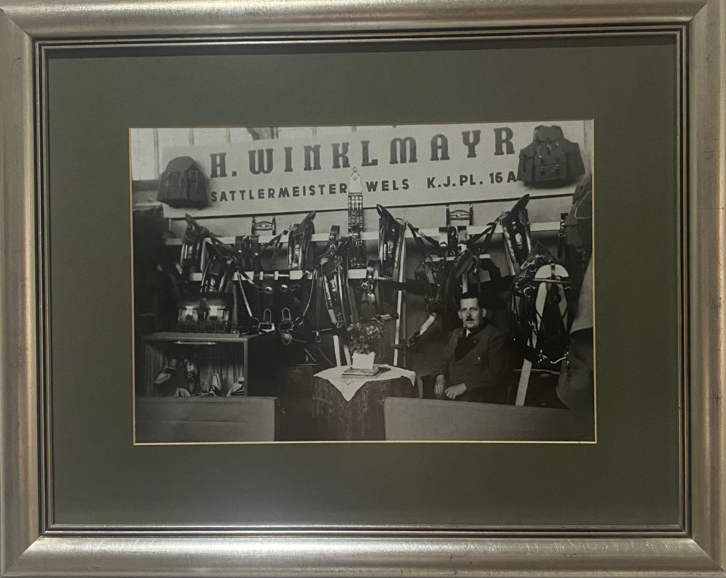 Historische Schwarzweißaufnahme: Sattlermeister H. Winklmayr vor seinem Stand mit Pferdegeschirr, Schild Sattlermeister Wels K.J.Pl. 16A