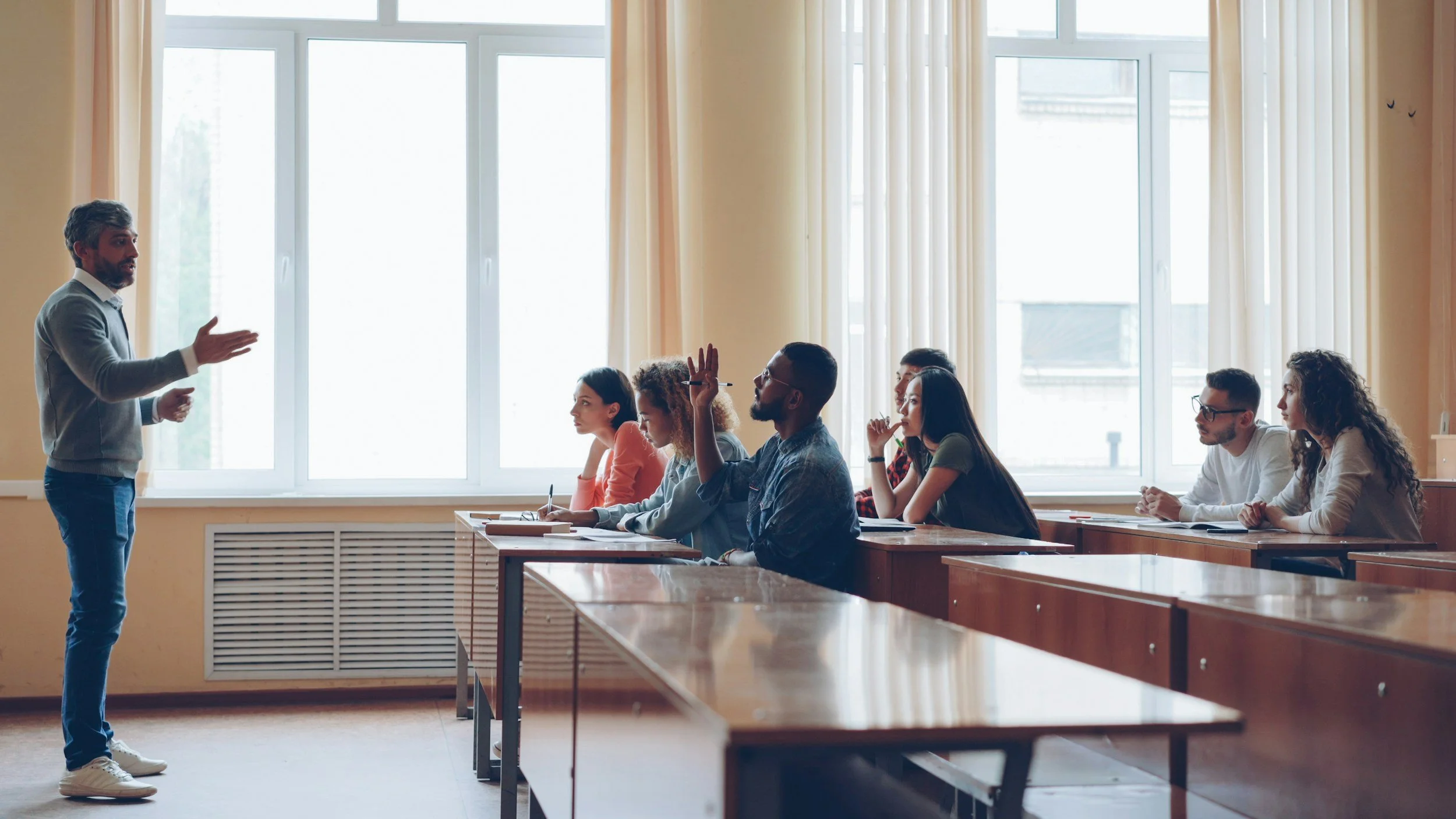 A classroom with a male teacher standing at the front and gesturing, while six students sit at desks listening. The classroom has large windows with curtains, and students are attentive, with one raising a hand.