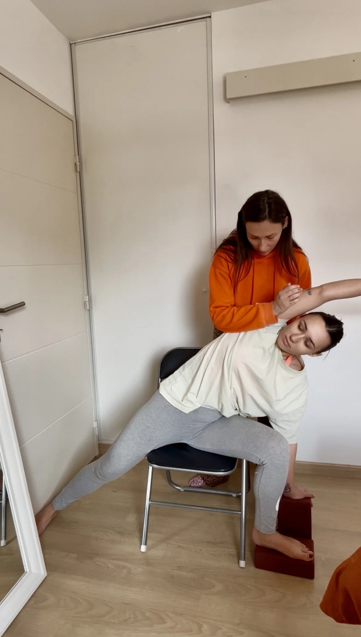 Une femme en position de stretching sur une chaise, une autre femme appuyée sur son bras en train de faire une manipulation. La scène se déroule dans une pièce intérieure avec un sol en bois et un mur blanc.