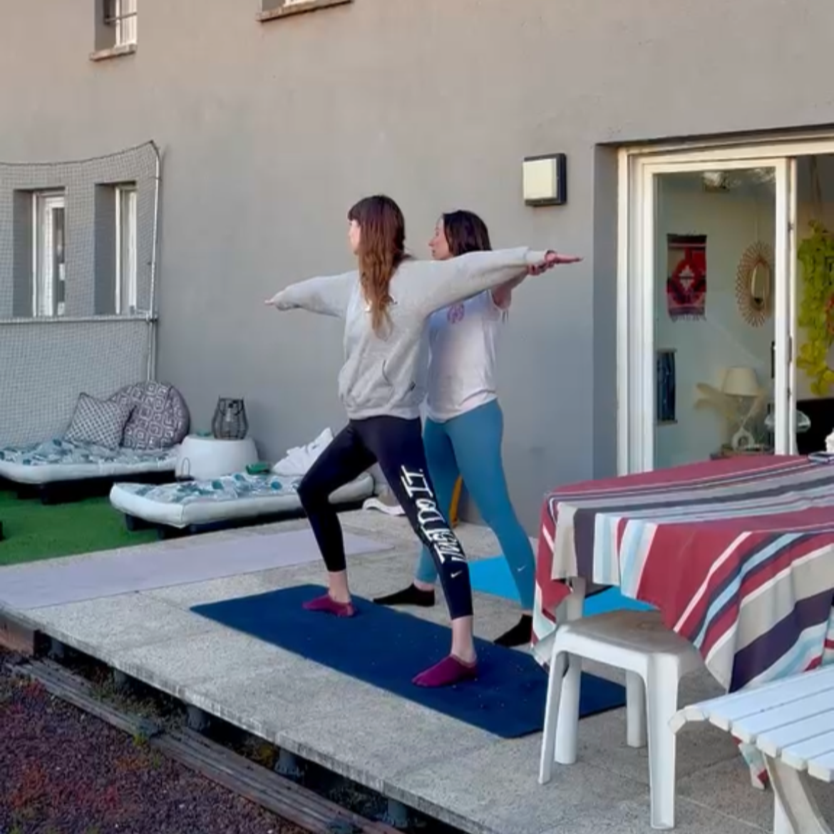 Deux femmes faisant du yoga en extérieur sur un tapis, face à une maison, avec des chaises et une table à côté.