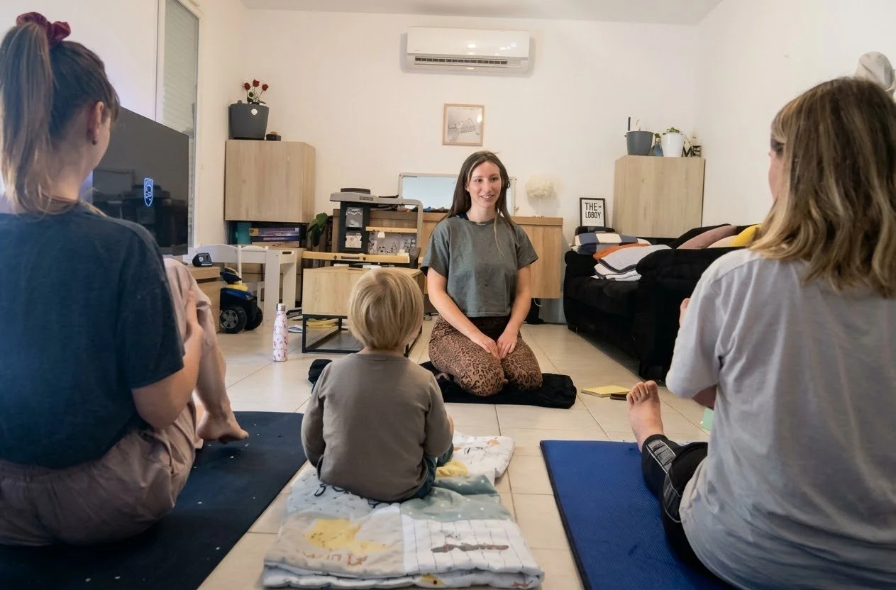 Un groupe de personnes, y compris un enfant, assis sur le sol d'une pièce, avec une femme assise en face, participant à une séance de groupe ou une activité de méditation dans une salle de salon.