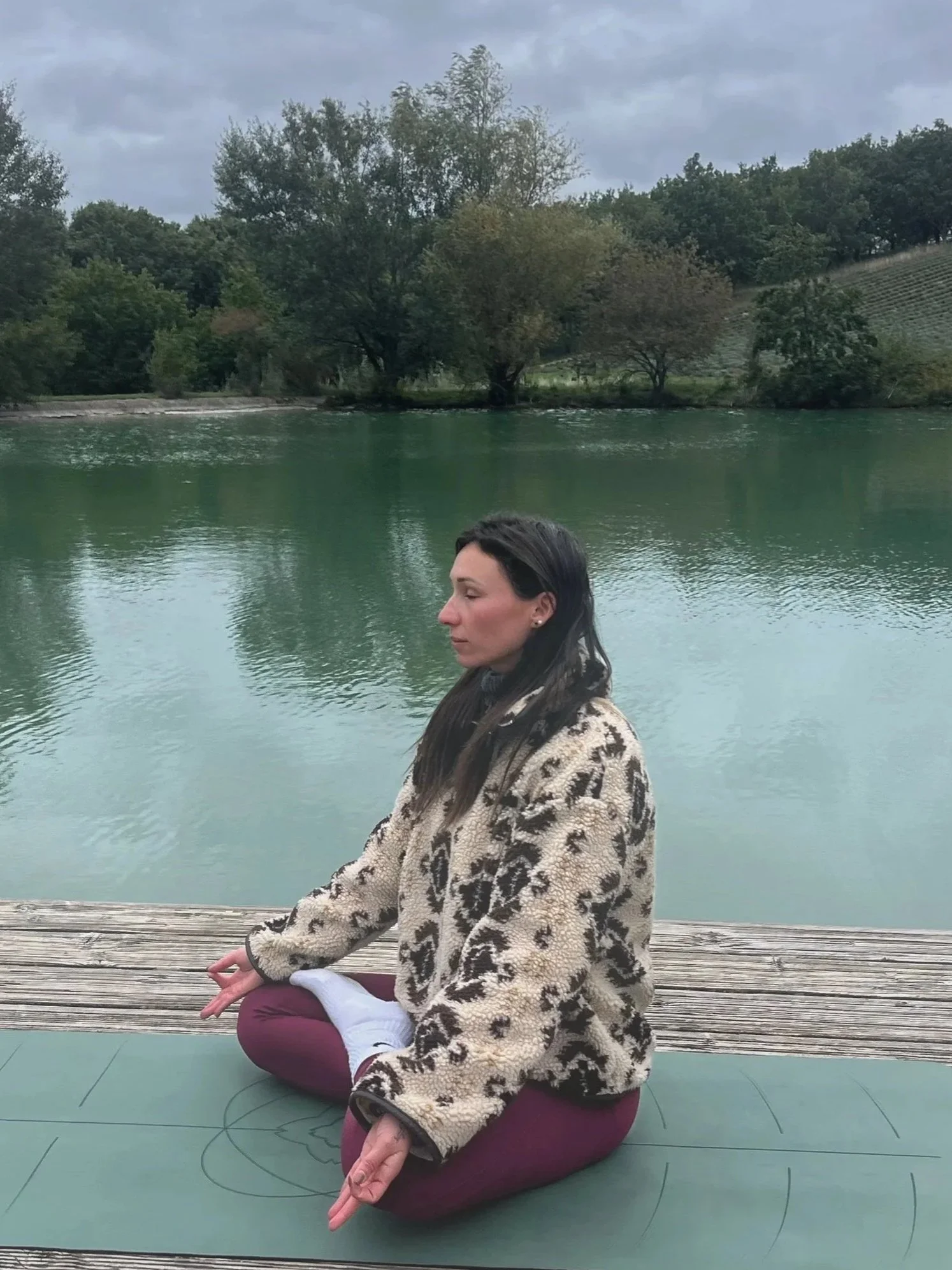 Une femme méditant en position du lotus près d'un lac, portant un manteau en fourrure et des pantalons bordeaux, avec des arbres et des montagnes en arrière-plan.