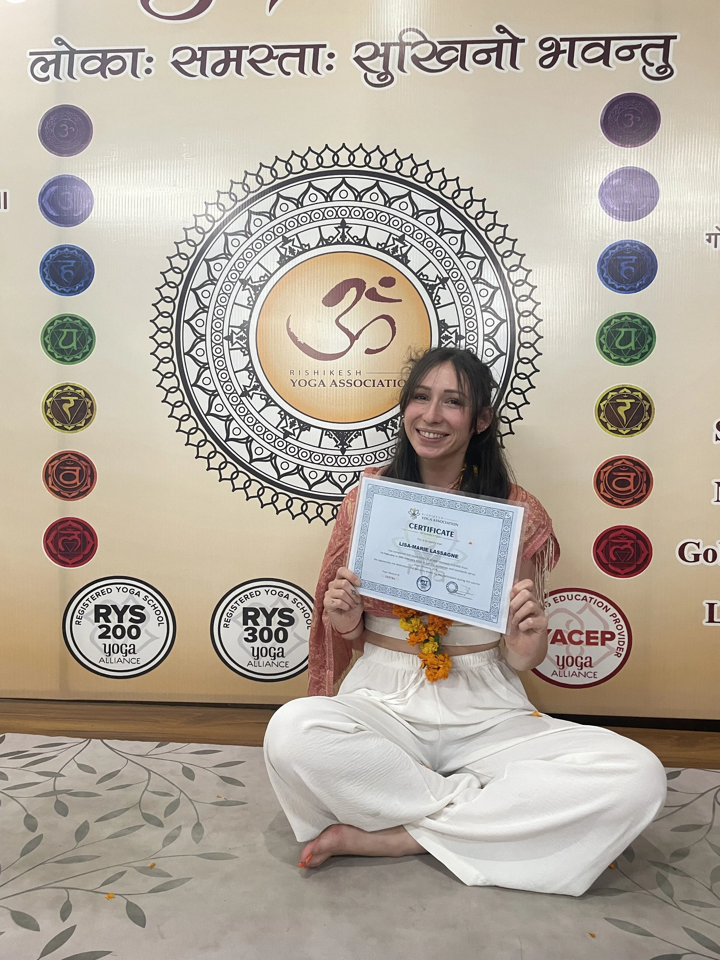 Femme souriante assise en position de méditation, tenant un certificat de réussite, portant un garland de fleurs, dans une salle avec un grand symbole Om et logos liés au yoga sur un mur en arrière-plan.