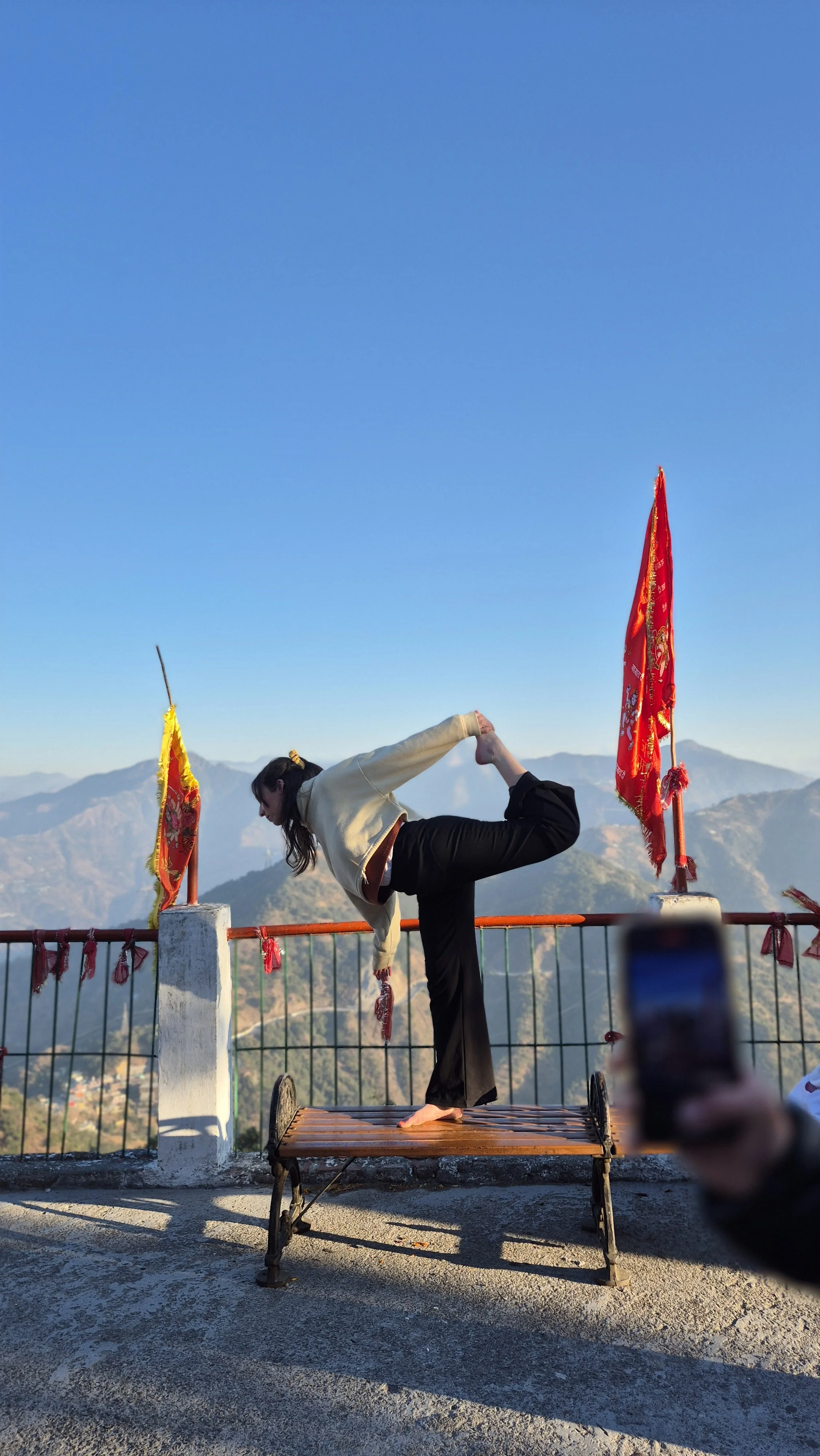 Une femme pratiquant le yoga en équilibre sur une patte sur un banc en bois, faisant une posture de yoga avec un paysage montagneux en arrière-plan, des drapeaux rouges et jaunes sur la balustrade environnante, sous un ciel bleu clair.