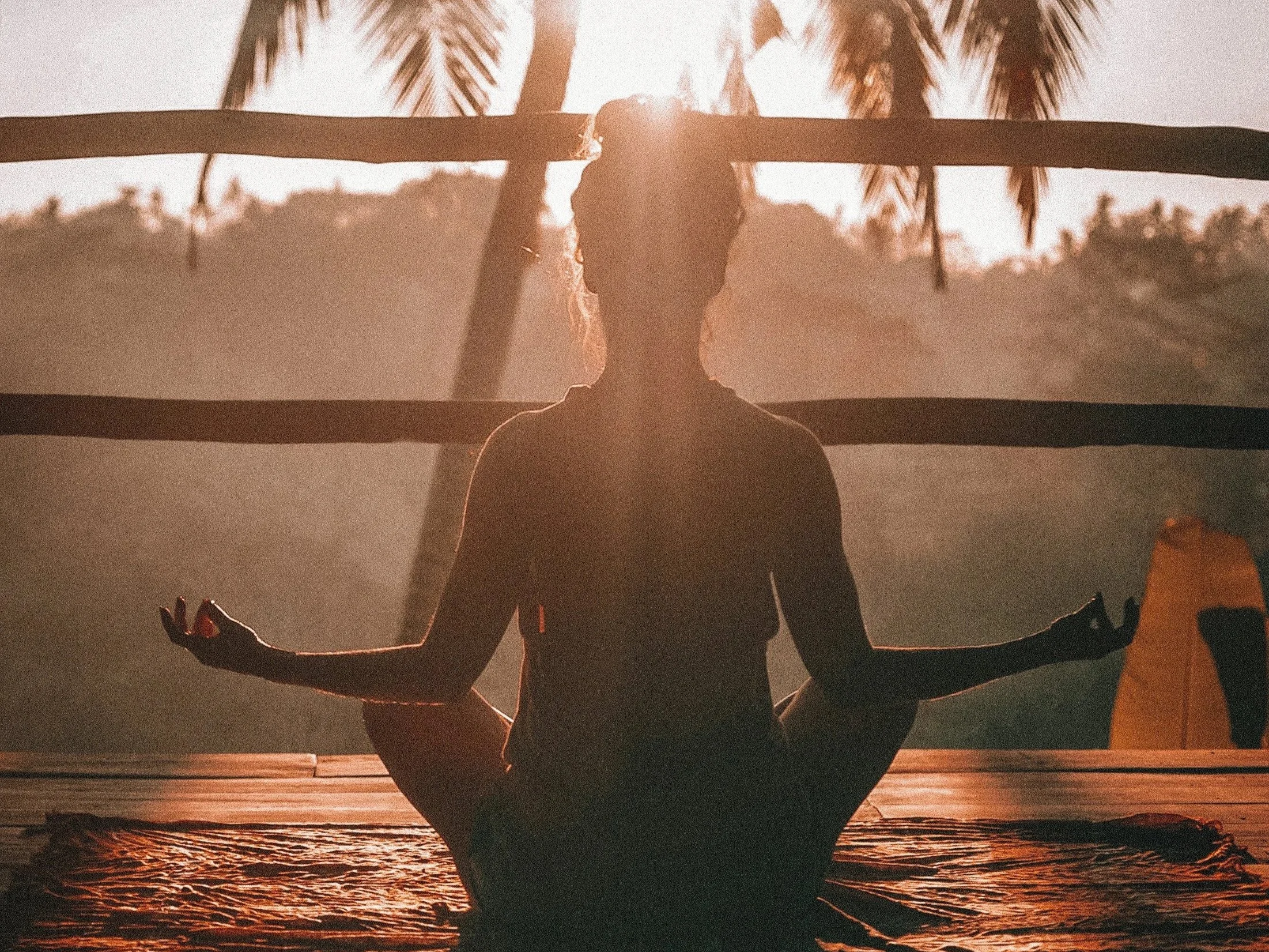 Silhouette d'une femme en position de yoga ou méditation sur une plage au coucher du soleil, avec des palmiers en arrière-plan.
