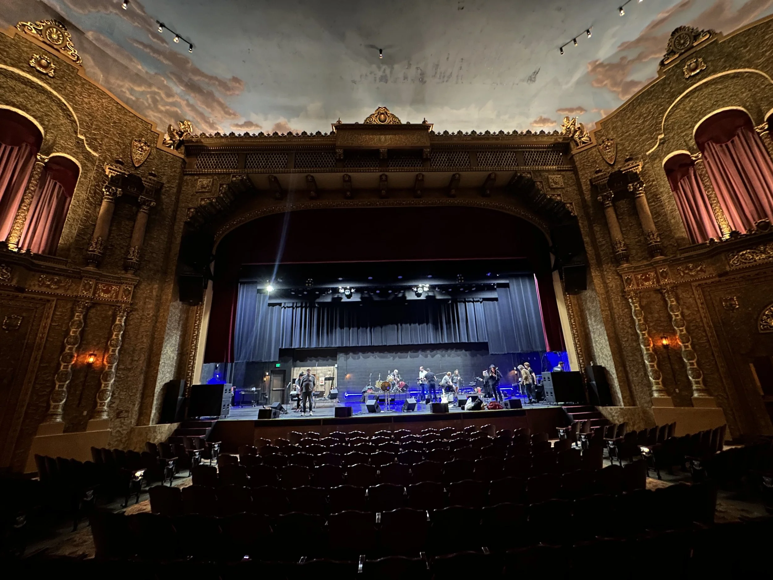 A theater stage set for a performance, with a painted ceiling depicting sky and clouds, ornate golden decorations, and red curtains on both sides. There are people on stage preparing, and empty audience seats in front.