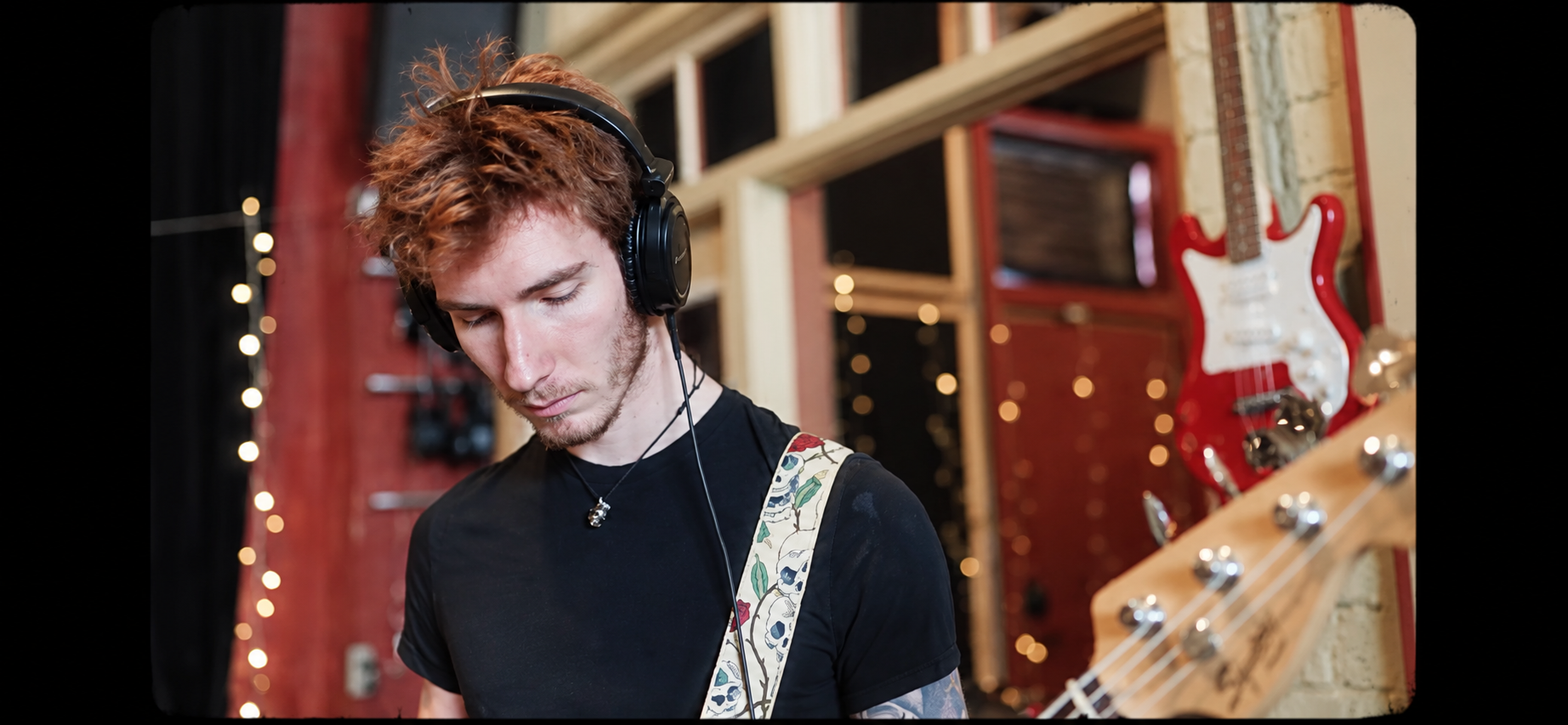 Young man with curly red hair wearing headphones, playing an electric guitar in a music studio decorated with string lights and featuring a red and white guitar on the wall.