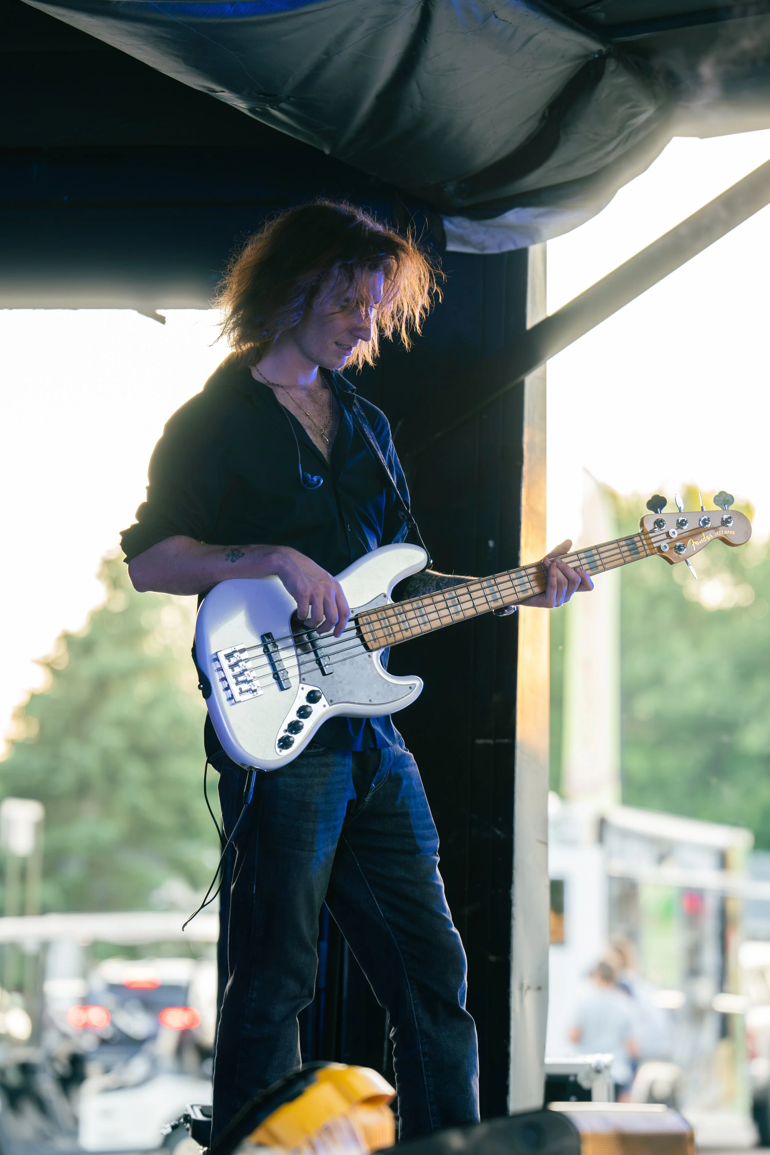 A musician playing a white electric bass guitar on stage during an outdoor performance at sunset, with a dark backdrop and natural light highlighting the musician.