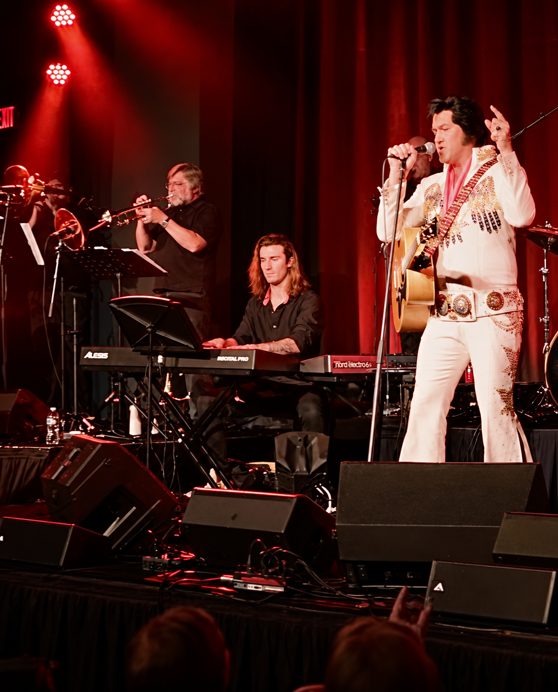 Elvis Presley impersonator singing into microphone with guitar on stage, accompanied by a keyboardist and a trumpet player in a concert setting with red lighting and stage equipment.