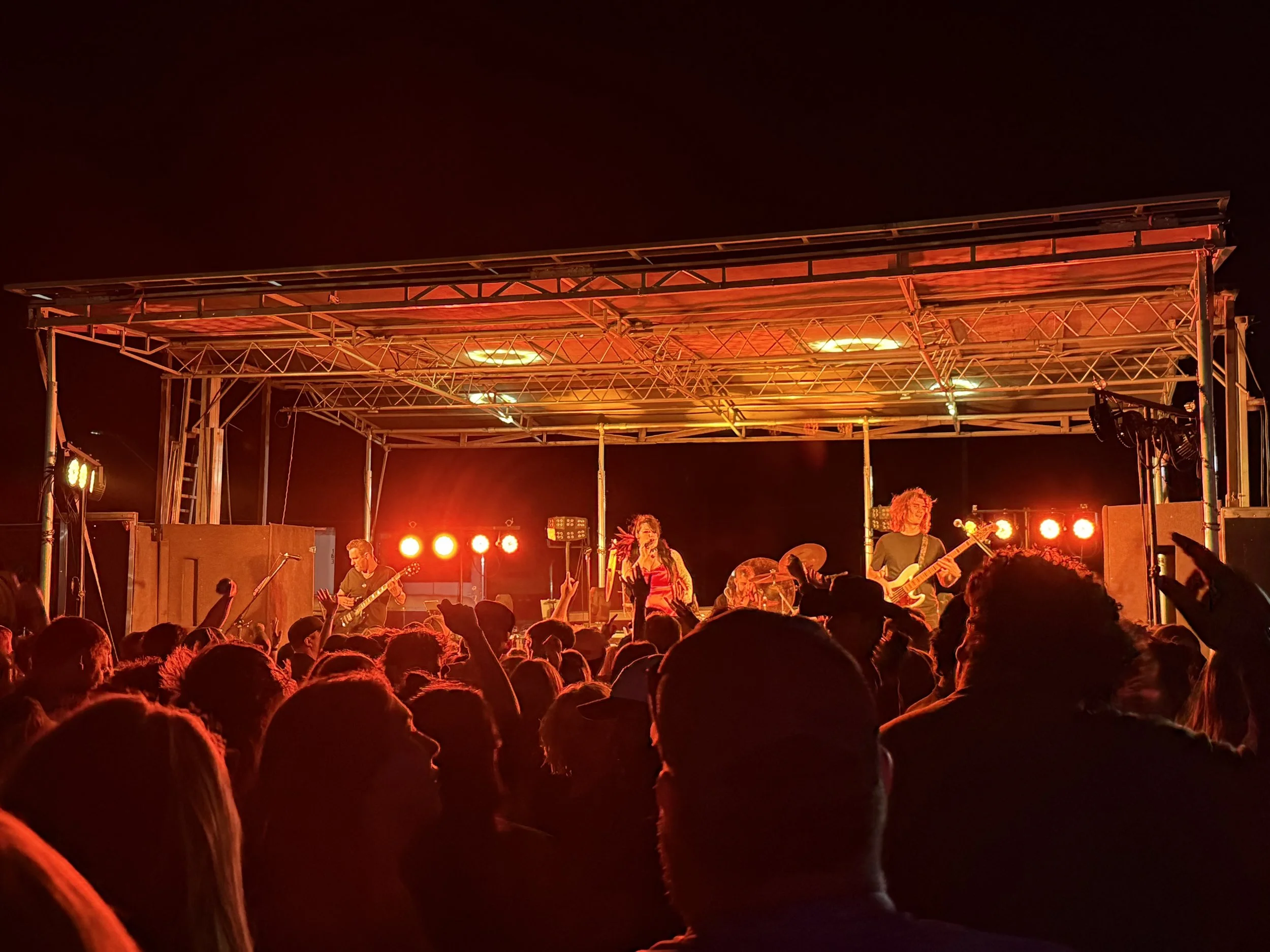 A live band performing on stage at a night outdoor concert. The audience is visible in the foreground, with many people raising their hands and enjoying the music.