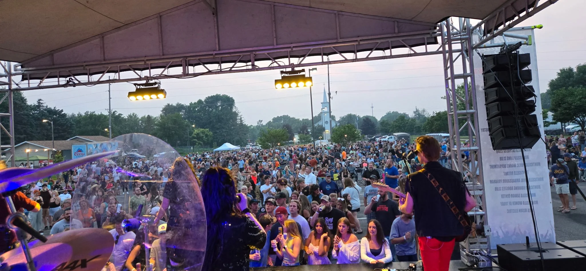 A large outdoor concert or festival with a crowd of people gathered in front of a stage. The image is taken from the stage, showing musicians performing. There are stage lights and large speakers visible, with a church steeple and trees in the backgr