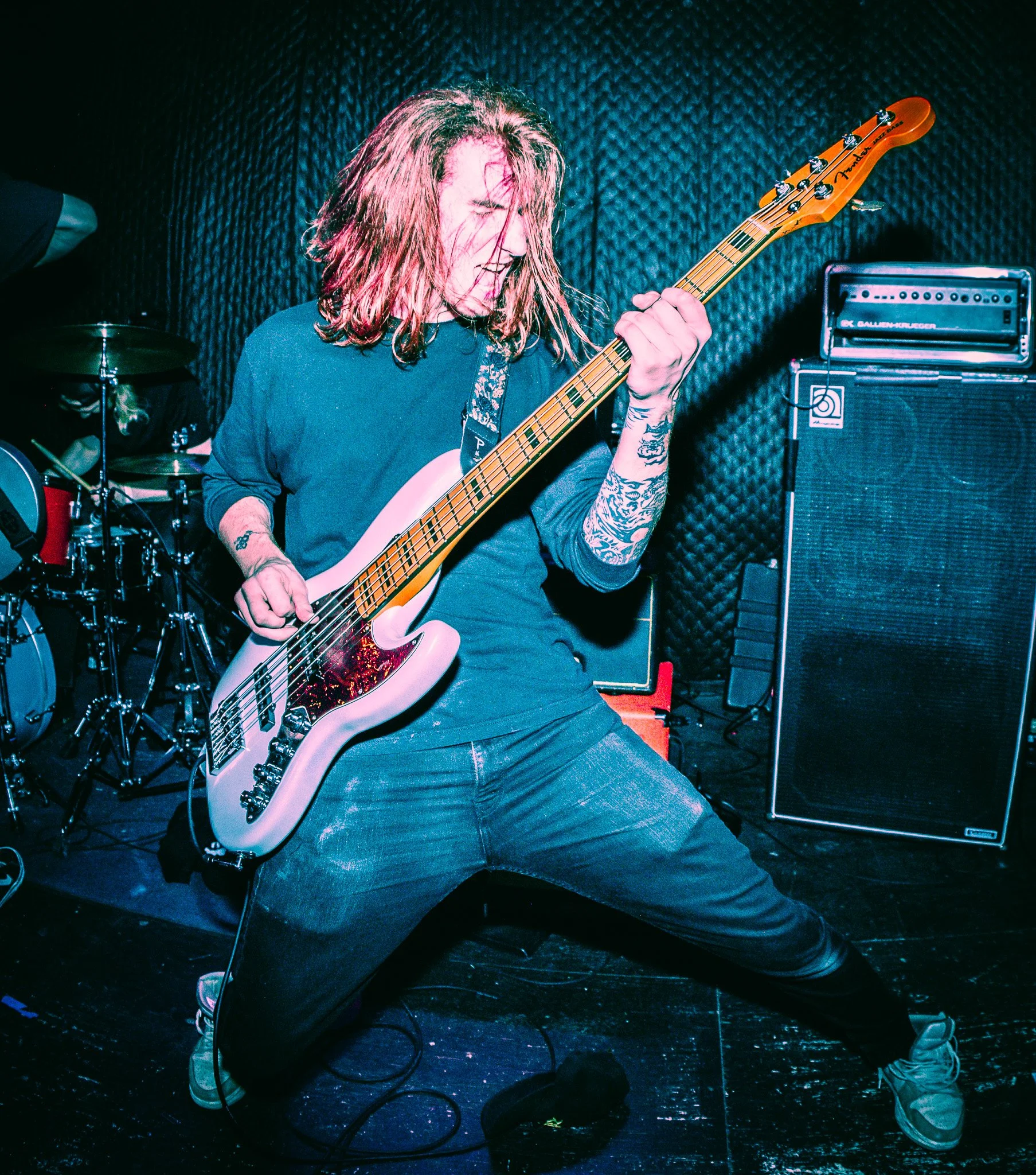 A musician with long hair playing a Fender bass guitar on stage, wearing dark jeans and sneakers, with a tattoo on his arm, in a music venue with black soundproof walls and musical equipment including a drum set and amplifier in the background.