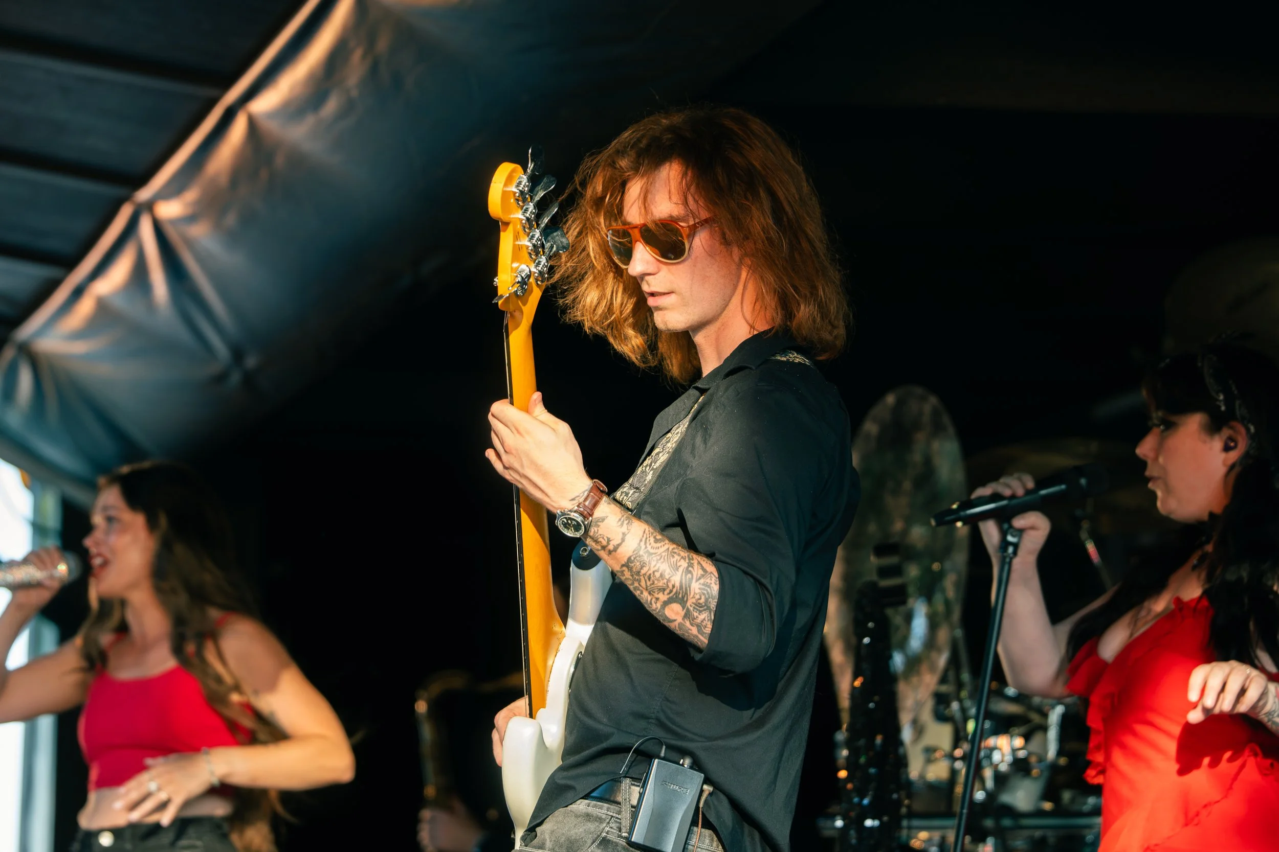 A musician with long hair, sunglasses, and tattoos is playing an electric guitar on stage during a concert. Several women with microphones are singing in the background.