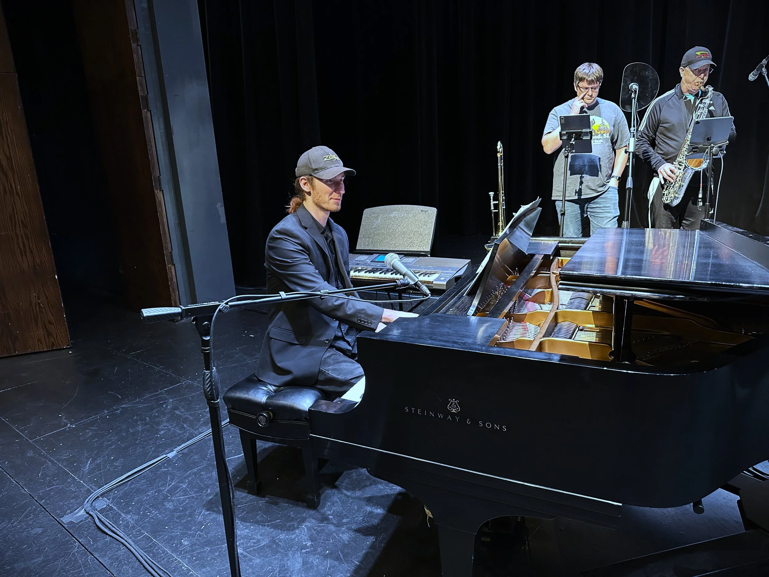 Three musicians performing on stage: one playing a grand piano, one holding a saxophone, and one with a music stand and tablet. The stage has a black background and wooden sides.