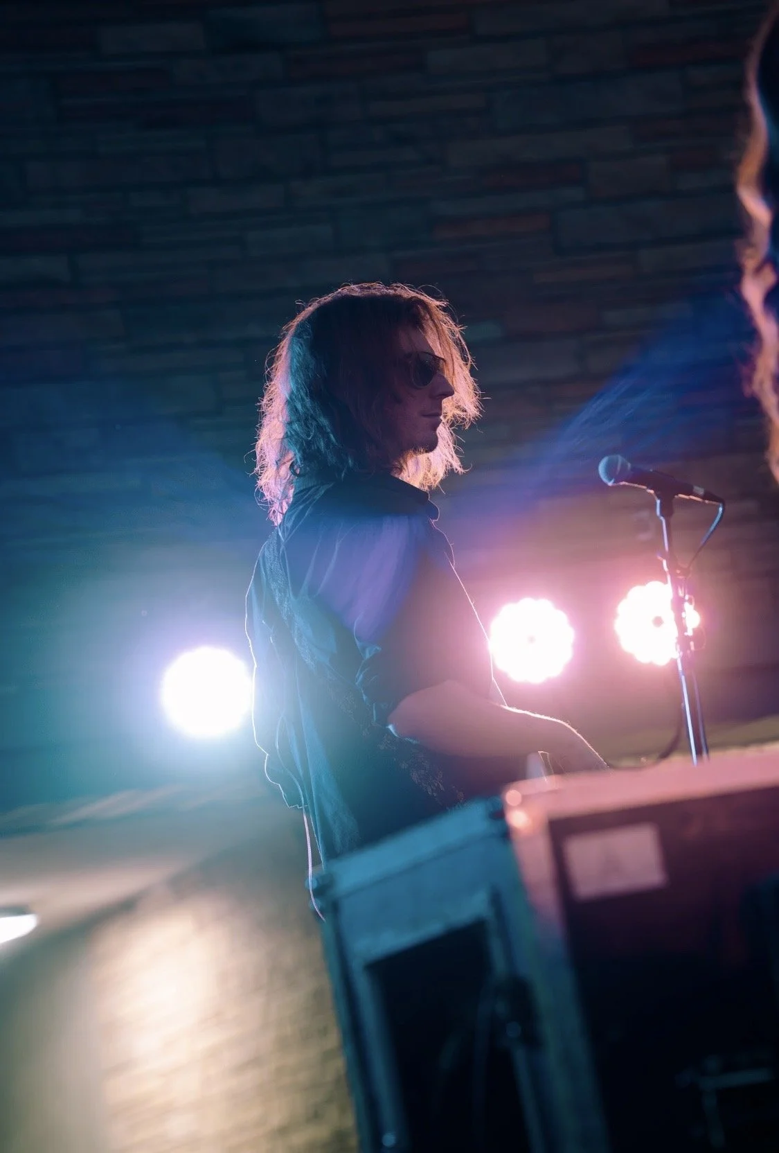 A person with shoulder-length hair and sunglasses playing a keyboard on stage with bright lights behind them.