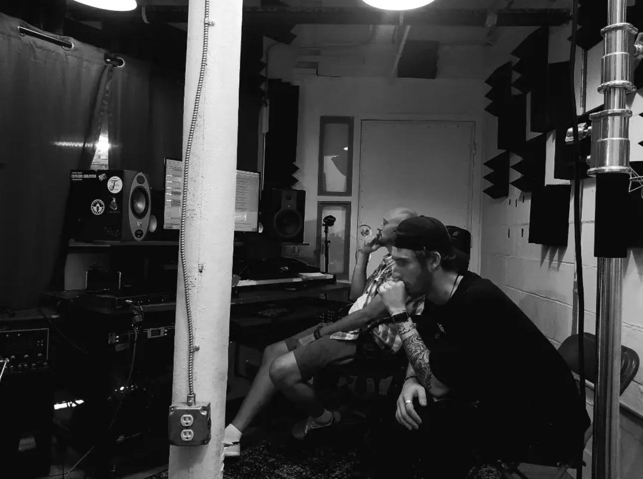 Two men sitting in a recording studio, listening to music or audio working with music equipment. The studio has acoustic foam panels on the walls and various audio gear.