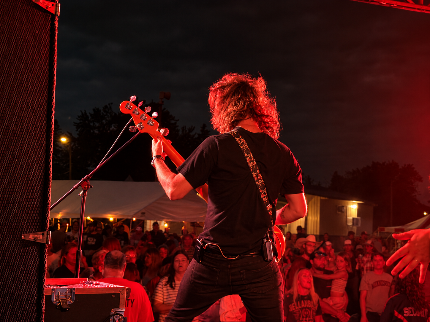 A guitarist performing at an outdoor concert at night, with an audience watching, lit by stage red lighting, and tents and trees in the background.