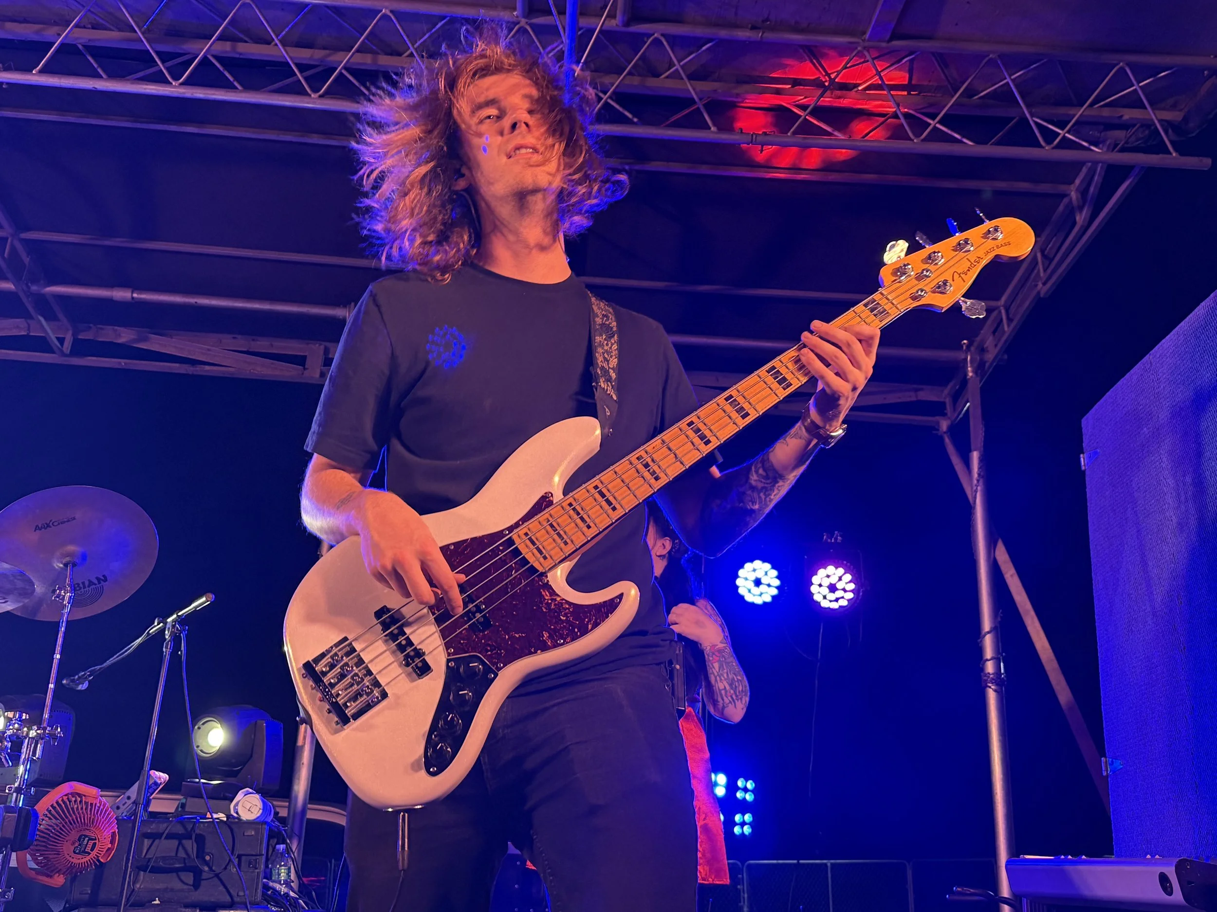 A musician with long curly hair playing a light-colored electric bass guitar on stage under purple and pink stage lights.