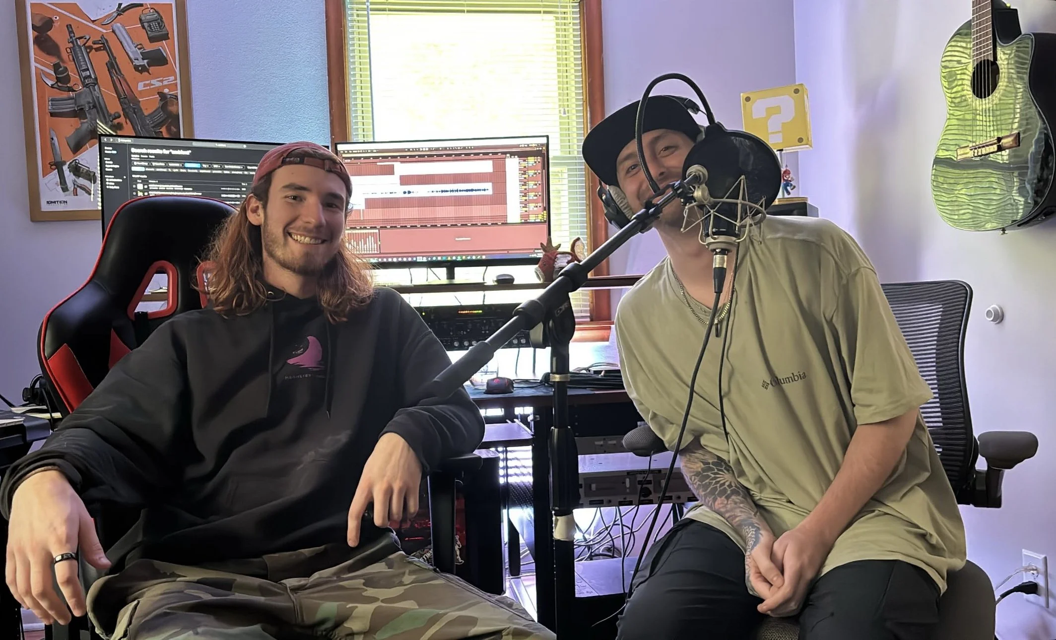 Two young men sitting in a home recording studio, smiling at the camera. One man has long hair, a baseball cap, and is wearing a black hoodie, while the other wears a black cap backwards, headphones, a beige Columbia shirt, and has tattoos on his arm. Behind them are computer monitors with digital audio workstations open, a guitar hanging on the wall, and a poster featuring various firearms.