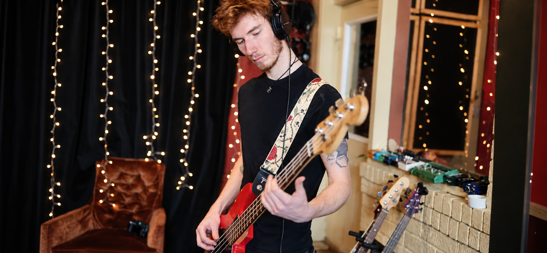 A young man with red hair, wearing a black shirt, headphones, and a patterned guitar strap, is playing a red electric bass guitar in a music studio with fairy lights in the background.
