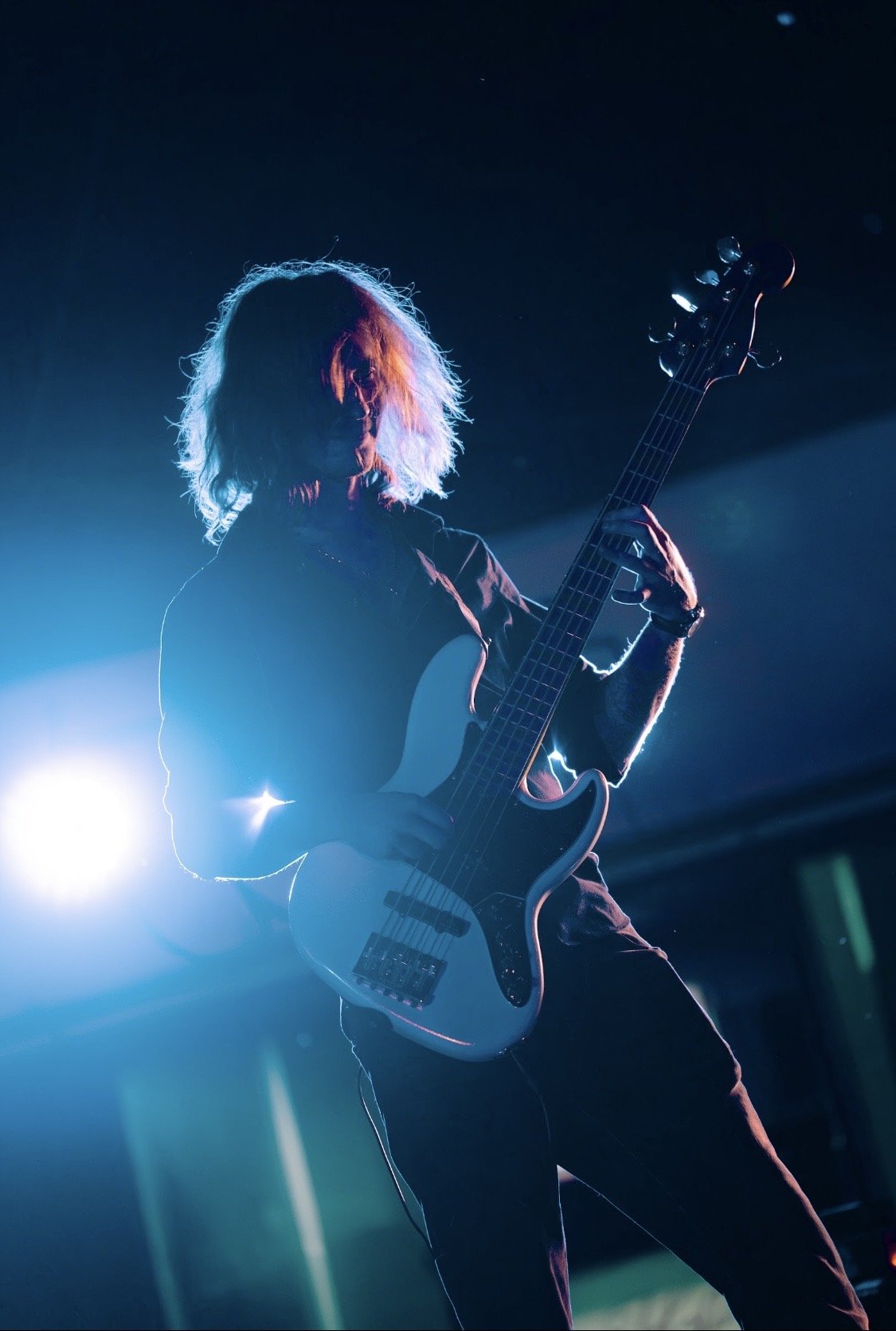A silhouette of a person with long hair playing an electric bass guitar on stage, backlit by bright blue and white lights.