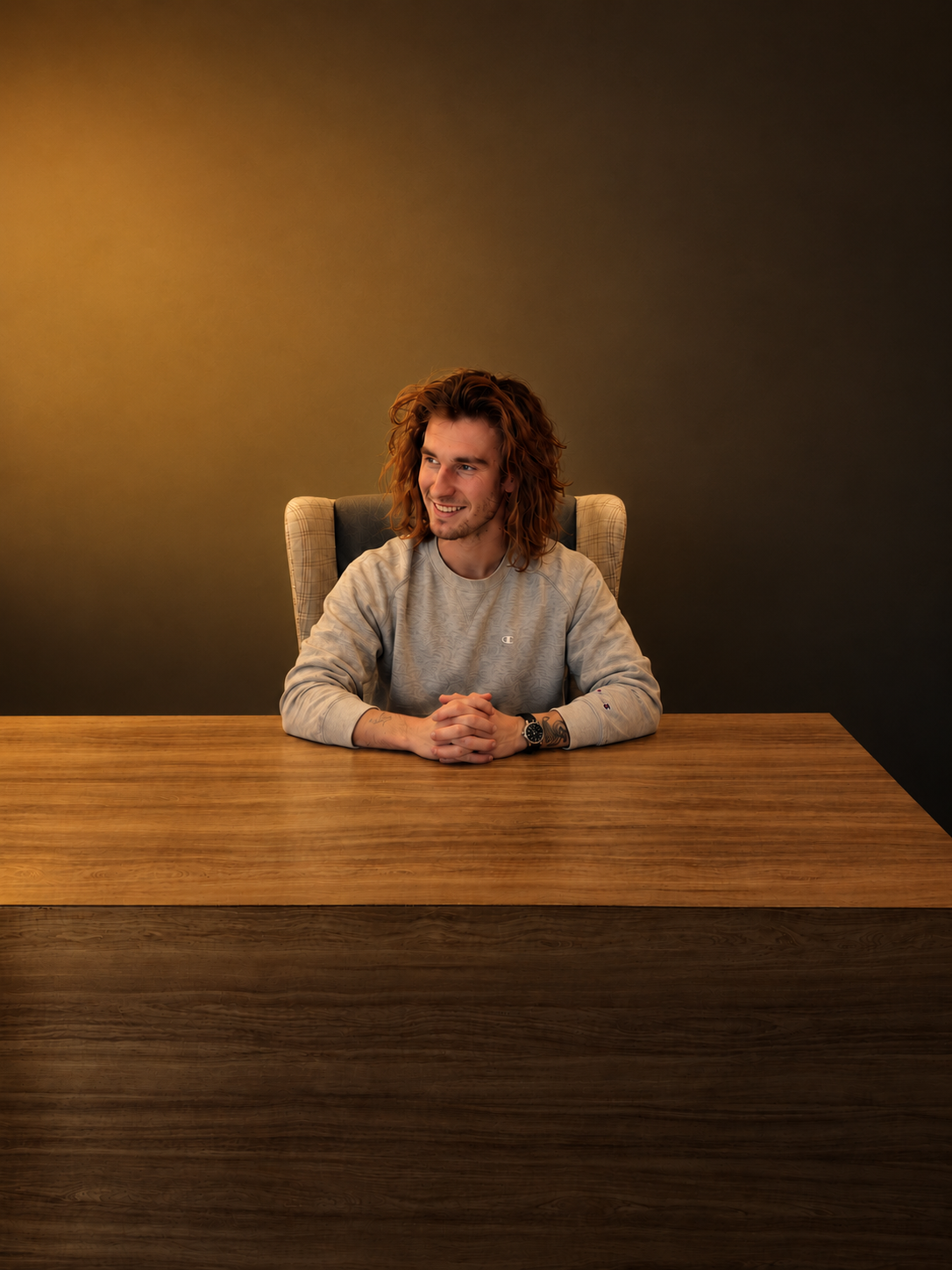 A young man with long, curly hair sitting at a wooden table with a dark background, smiling and looking to the side.
