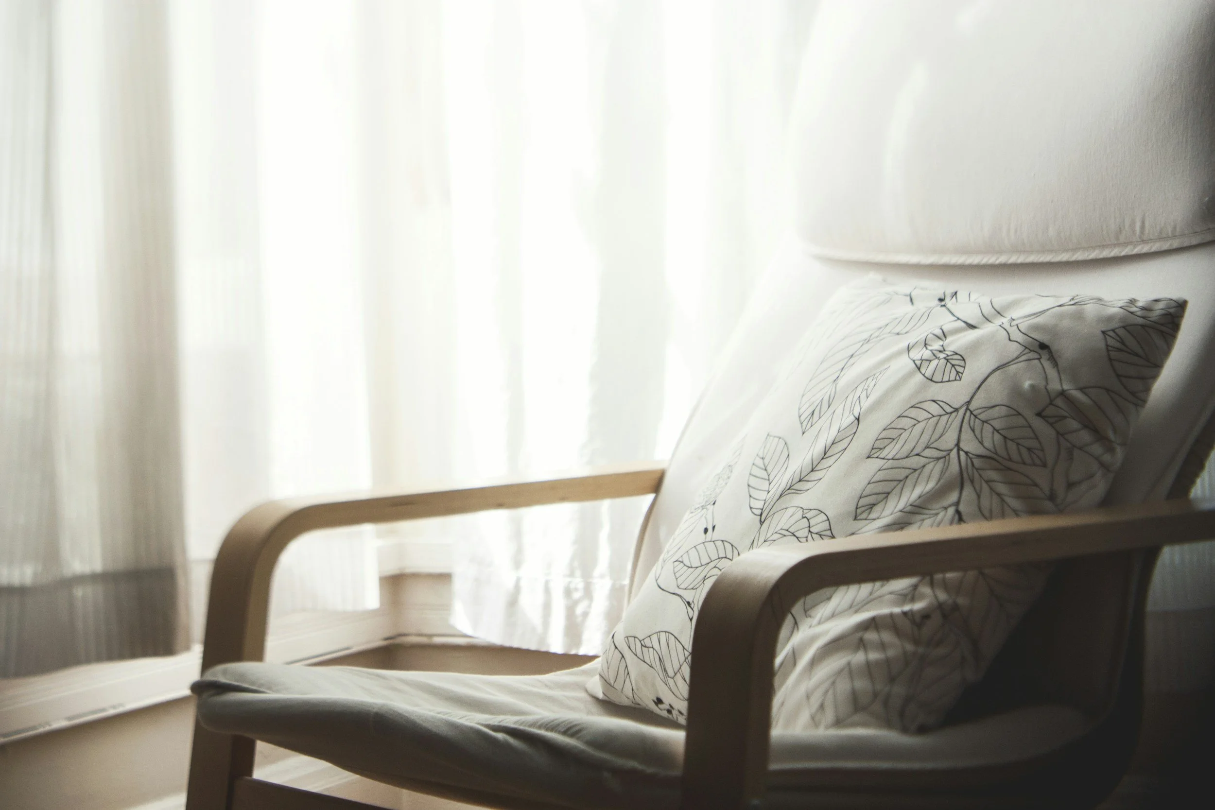 Cozy wooden armchair with gray cushion and patterned leaf-print pillow beside a sunlit window.