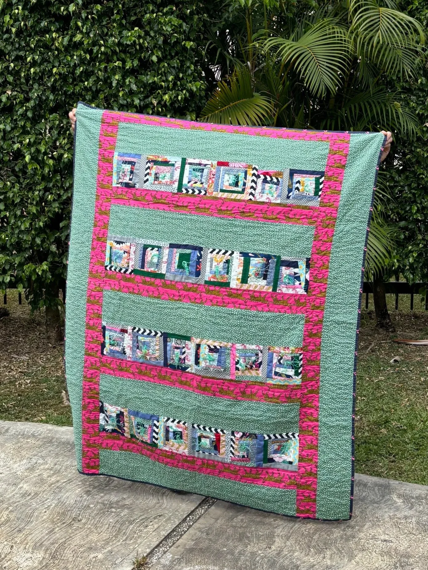 Handmade patchwork quilt with green, pink, and multicolored fabric strips displayed outdoors in a garden