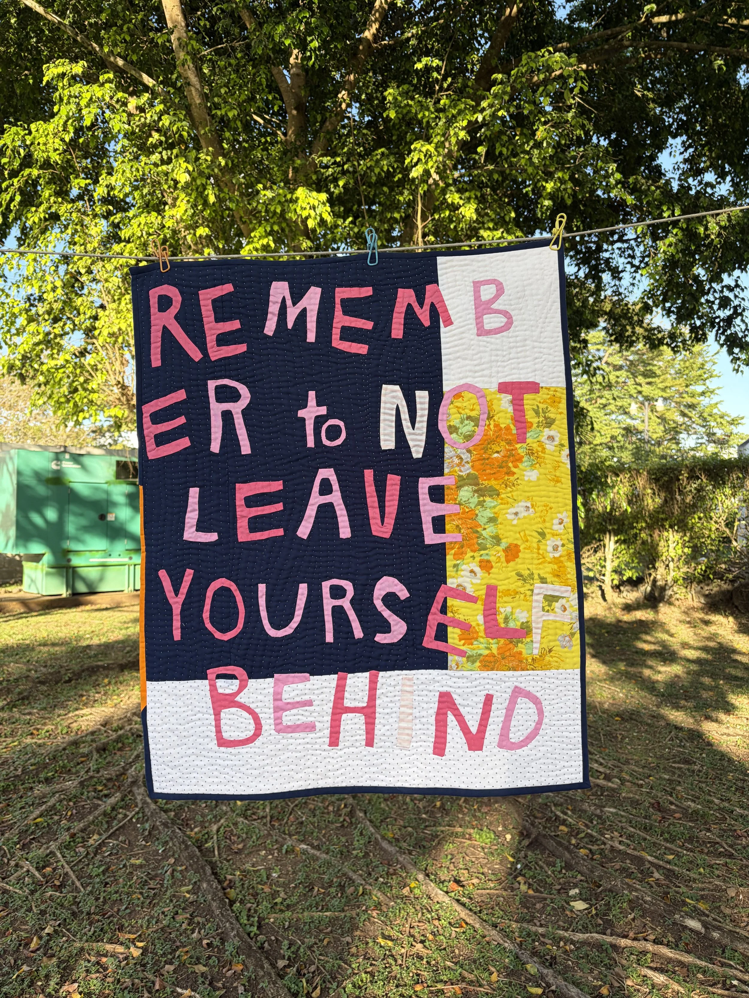 Handmade quilt sign hanging outdoors that reads “Remember Every Moment Yourself Be Kind,” with trees in the background.