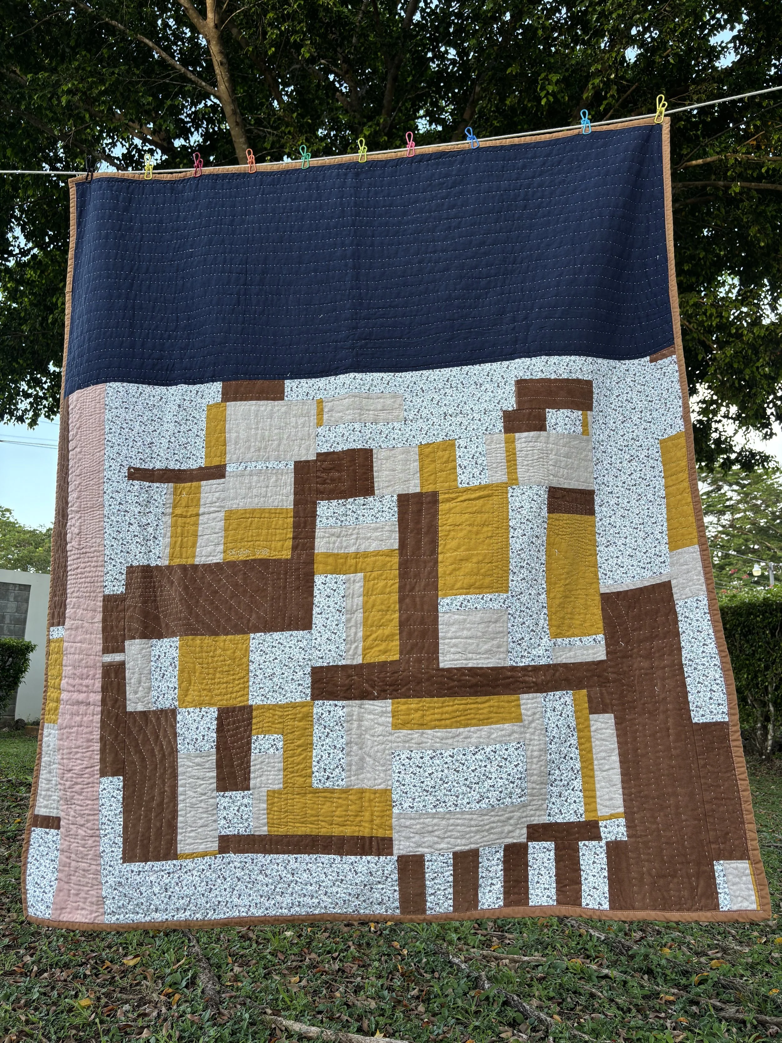 Large handmade patchwork quilt hanging outdoors on a clothesline with colorful clothespins, featuring navy, brown, mustard, gray, pink, and floral geometric fabric blocks.