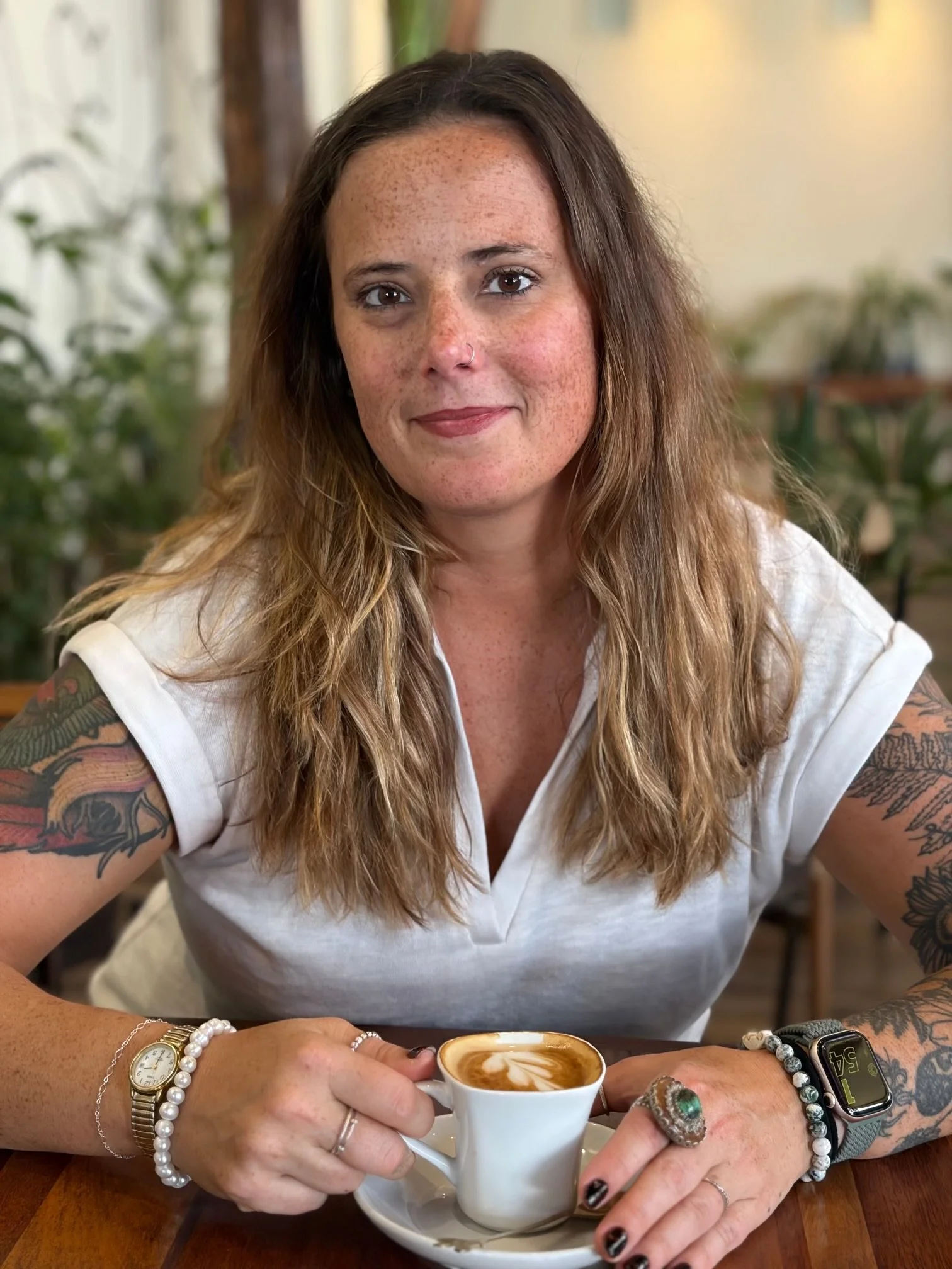Tattooed woman with long brown hair holding a cup of latte at a cafe table