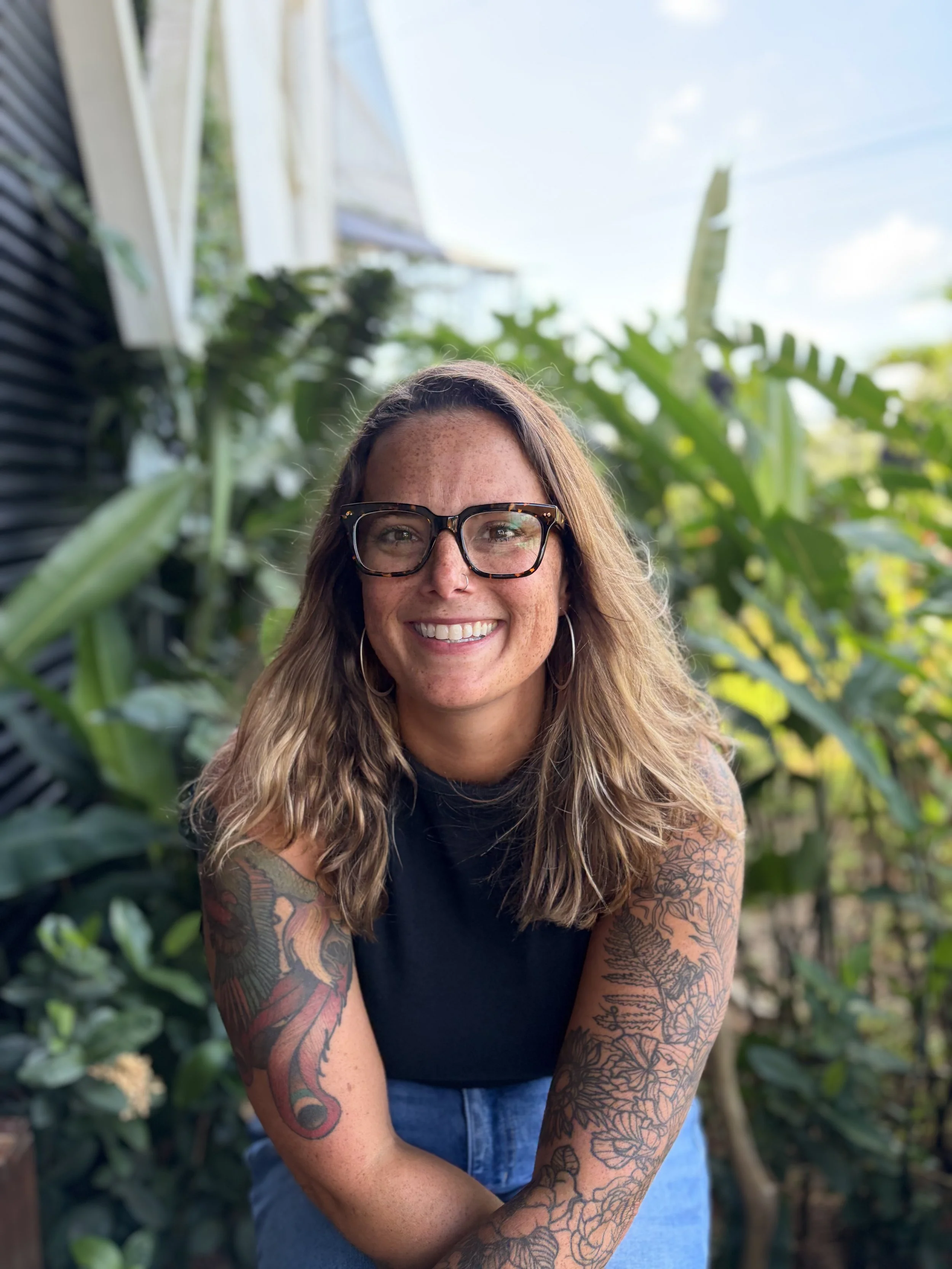 Smiling woman wearing glasses and a black sleeveless top, standing among lush tropical plants.