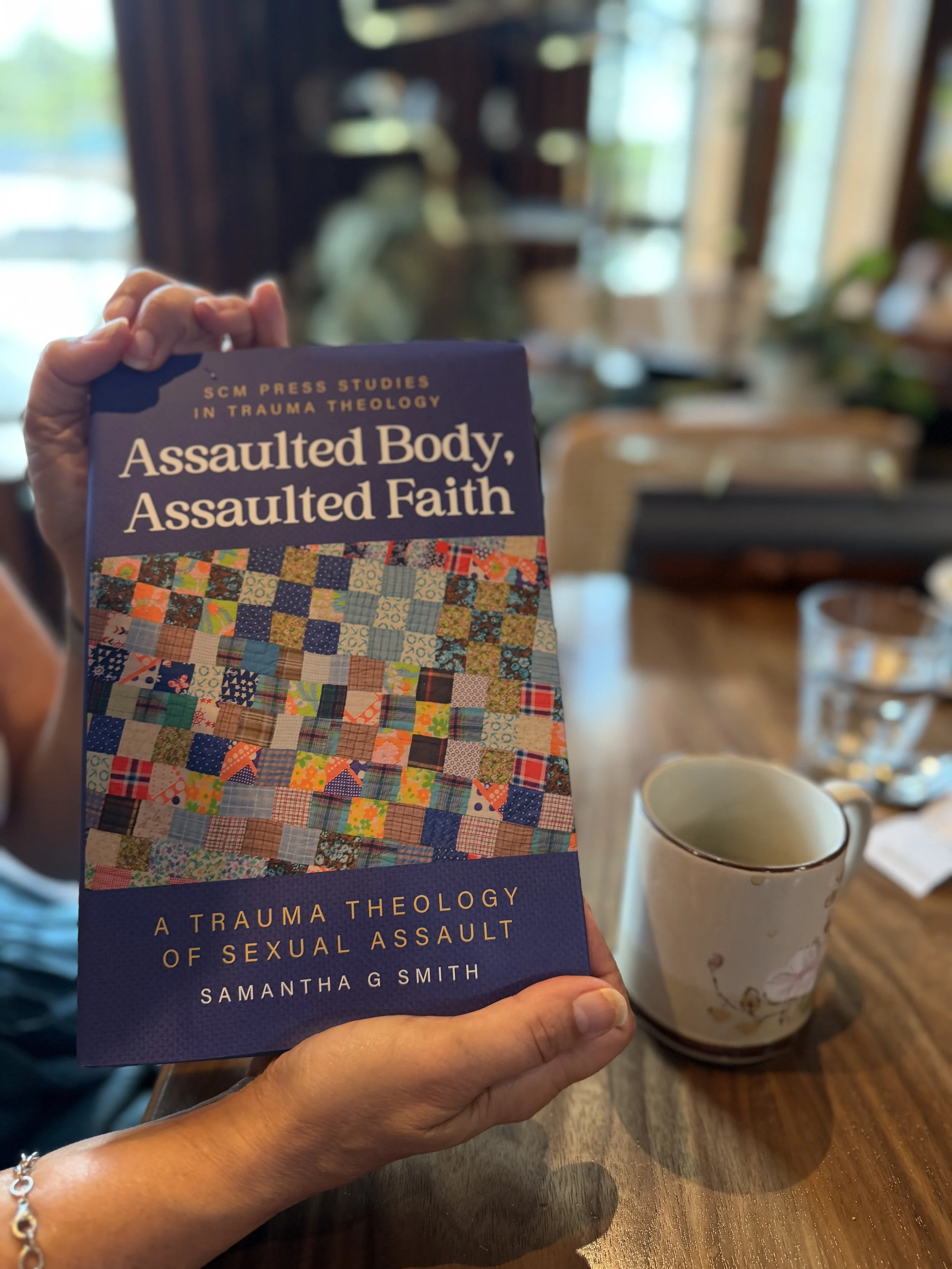 Hands hold the book “Assaulted Body, Assaulted Faith: A Trauma Theology of Sexual Assault” by Samantha G. Smith beside a coffee cup.
