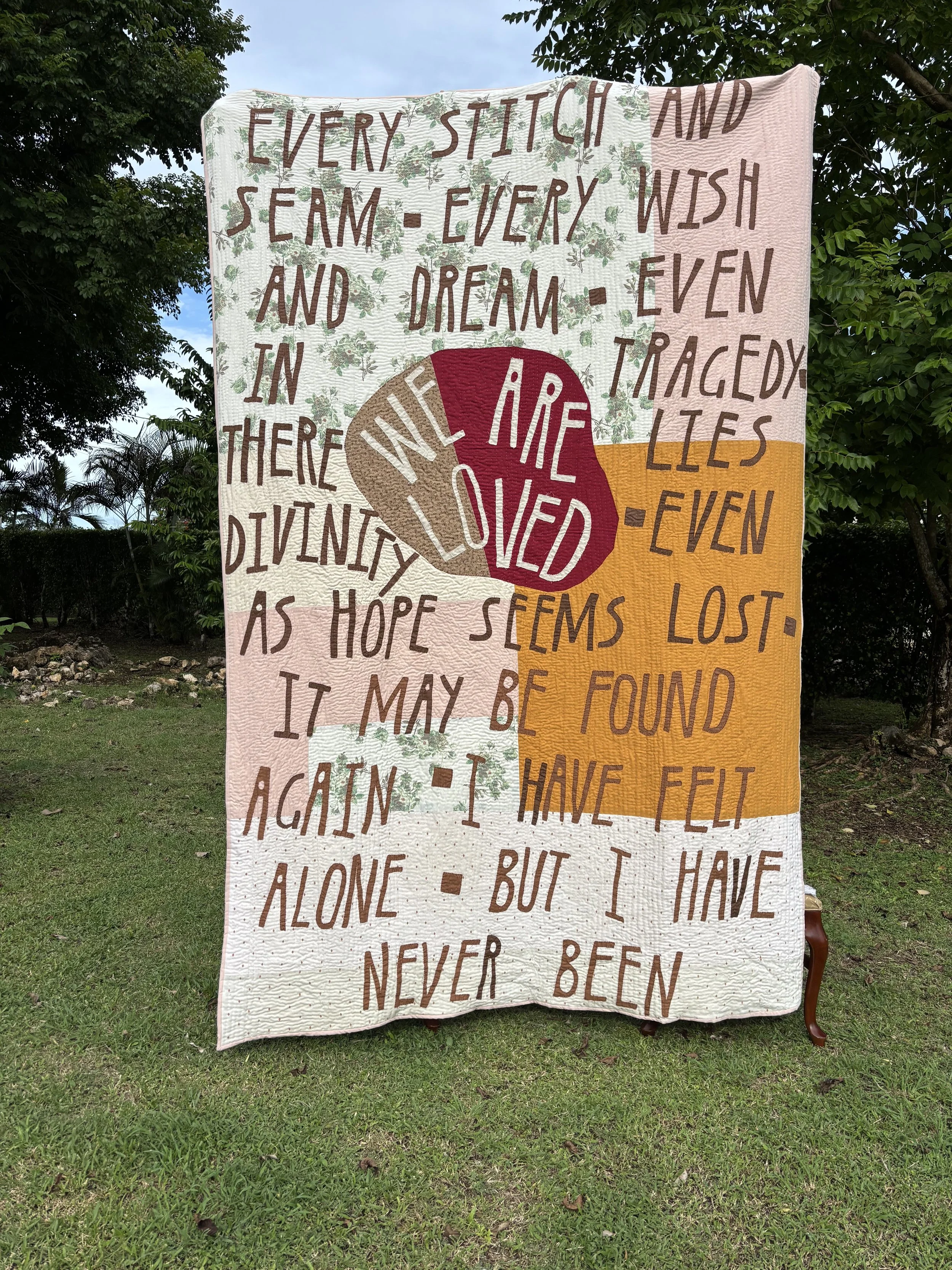Colorful quilt draped over a chair outdoors, featuring floral panels, bold text, and a central peace symbol.