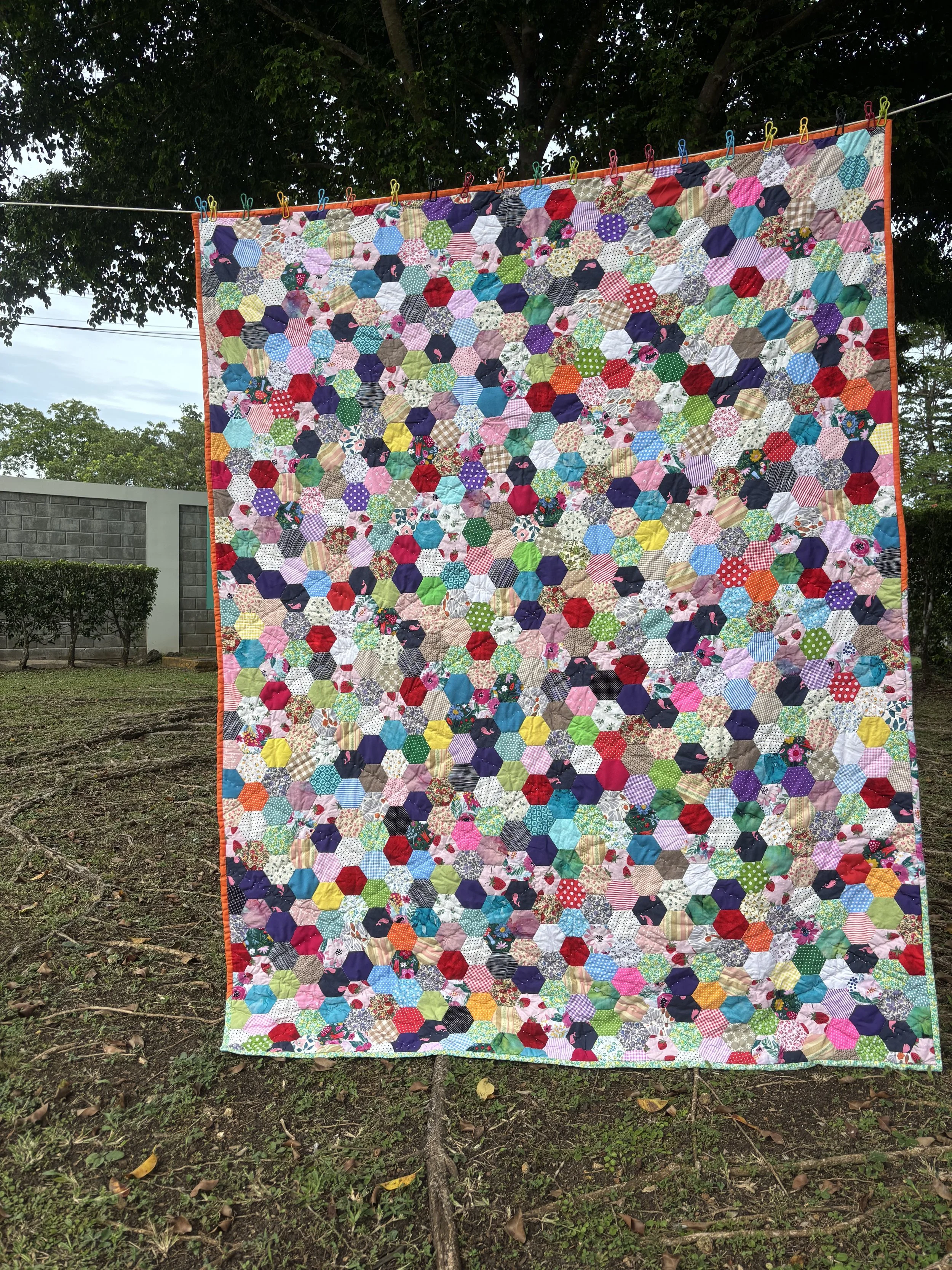 Colorful hexagon patchwork quilt hanging outdoors on a clothesline with clothespins, surrounded by trees.
