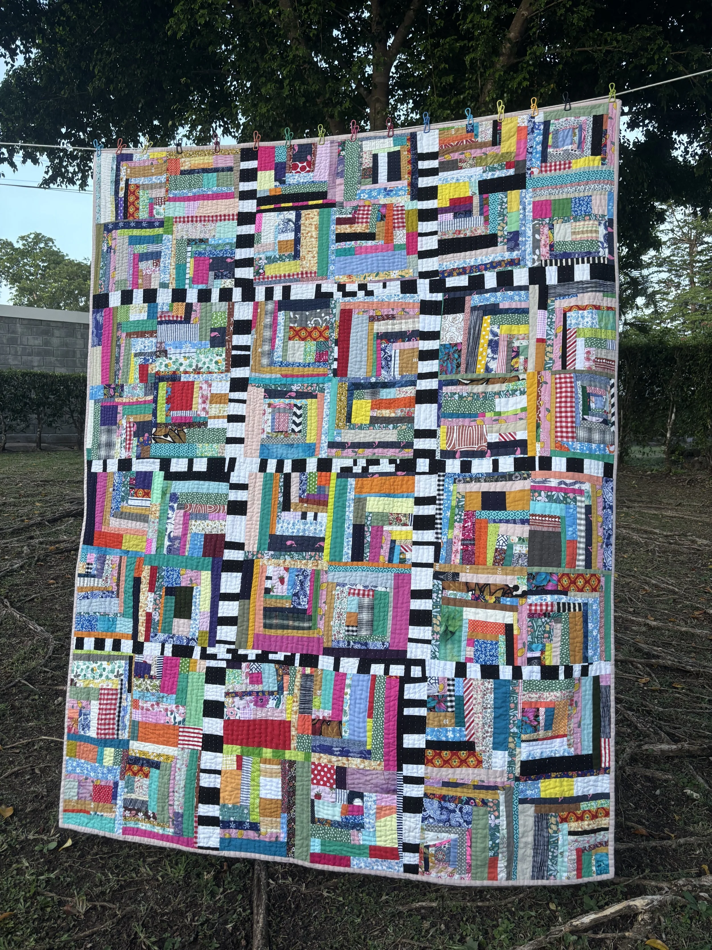 Colorful handmade patchwork quilt hanging outdoors on a clothesline, secured with clothespins beneath trees.
