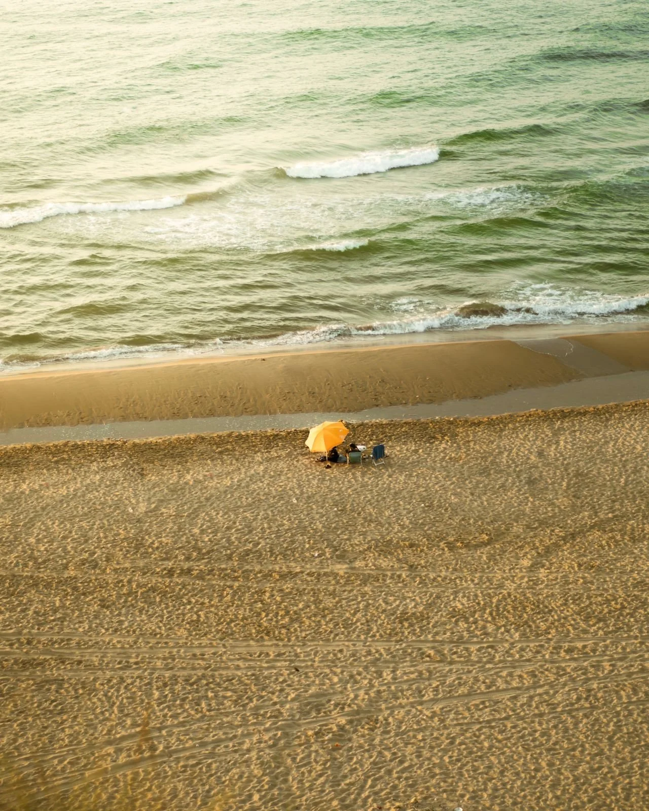 Empty beach with a yellow umbrella and two chairs near the shoreline, overlooking the ocean with small waves.