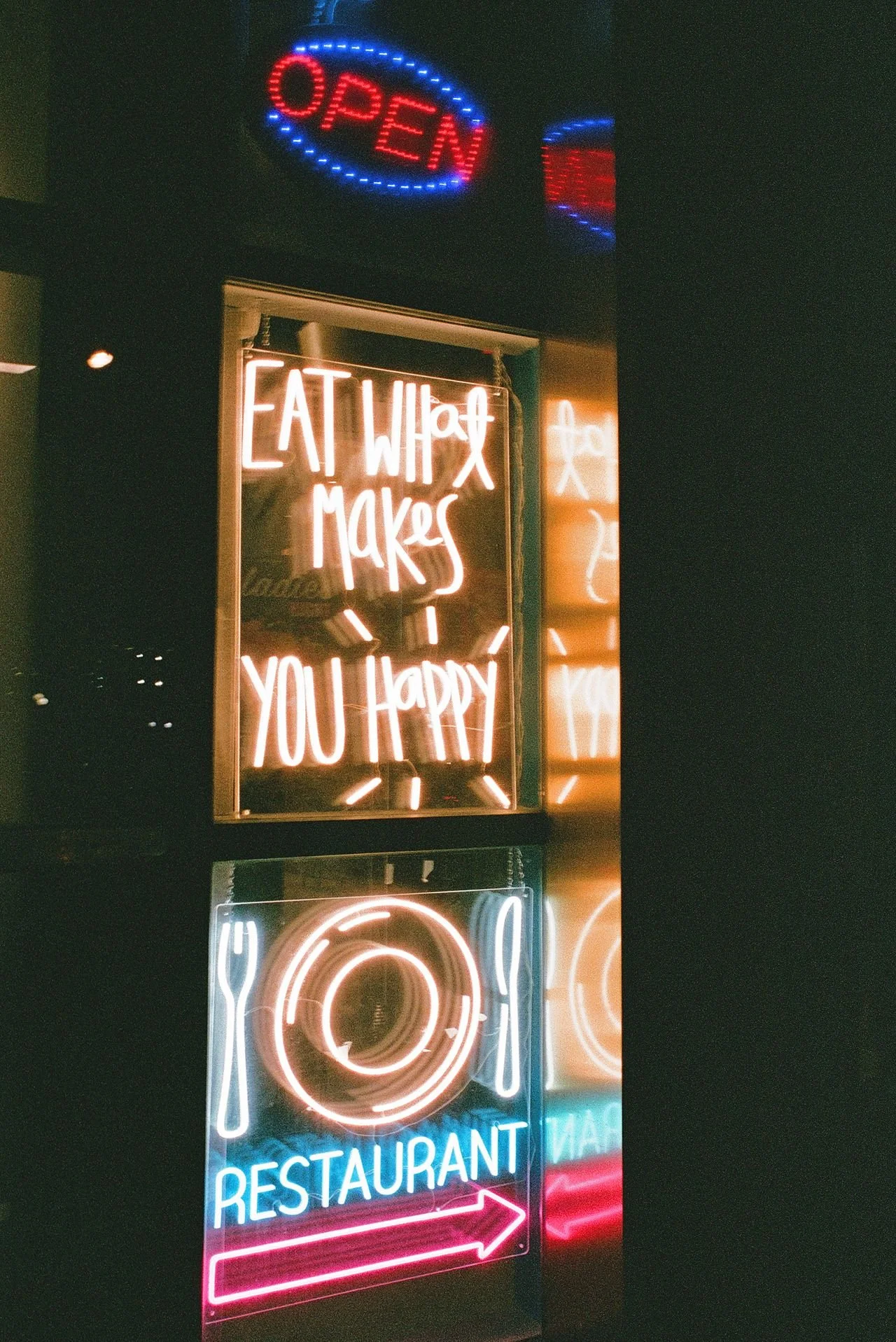 illuminated neon signs outside a restaurant. The signs display phrases like 'Eat What Makes You Happy,' an 'Open' sign, and an arrow pointing to the right.