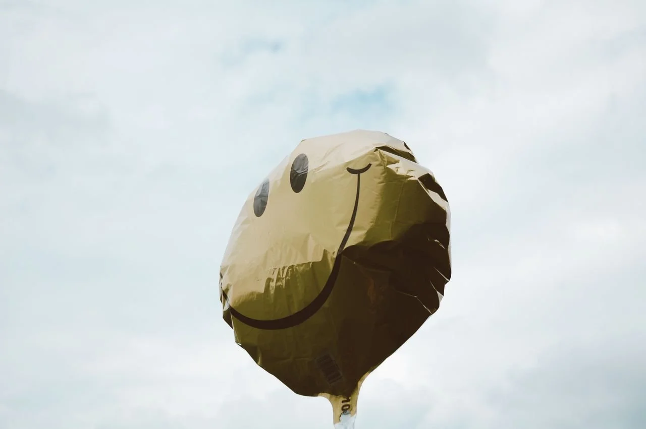 A yellow balloon with a smiley face design, floating in the sky with clouds.