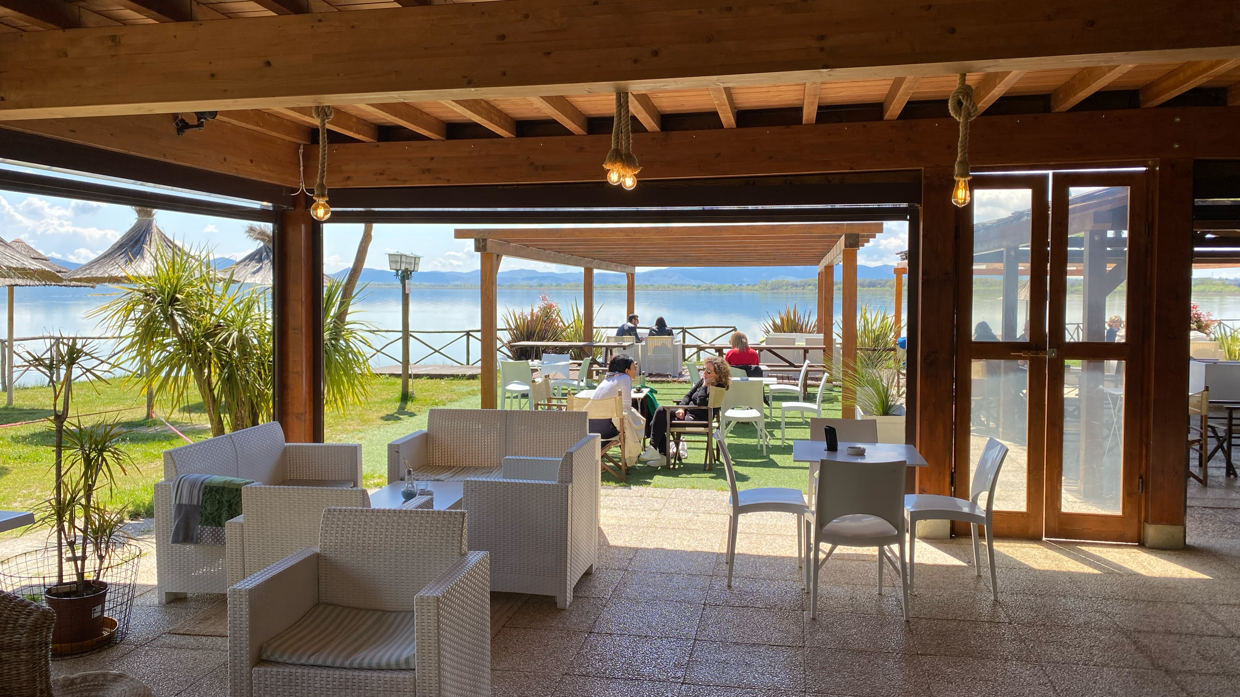 Indoor view of a lakeside outdoor dining area with wooden ceiling, hanging light bulbs, and a large window opening to a grassy area with outdoor seating. Beyond the grass, there is a lake with a clear sky and mountain range in the distance, with seve