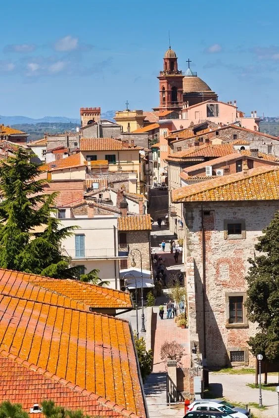 A view of Castiglione Del Lago with narrow streets, colorful buildings with orange tiled roofs, and a church with a bell tower in the background under a blue sky.