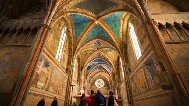 Interior of a historic Assisi cathedral with ornate vaulted ceilings, stained glass windows, and religious murals on the walls, with people walking inside.