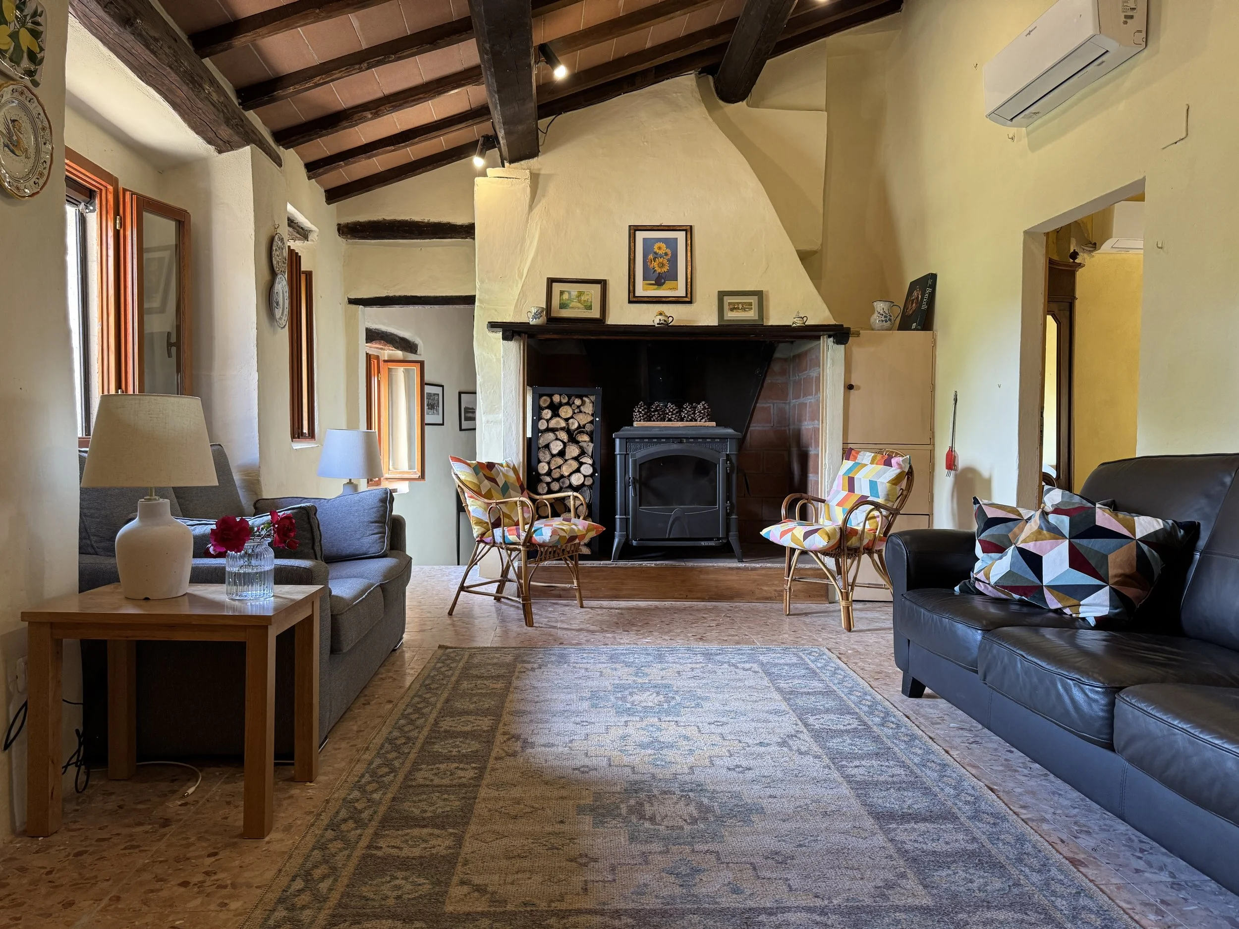 A cozy living room with a wood-burning stove, wood beamed ceiling, a gray sofa, and various framed artwork on the walls. Multiple windows with wooden frames let in natural light. There is a fireplace with stacked firewood.