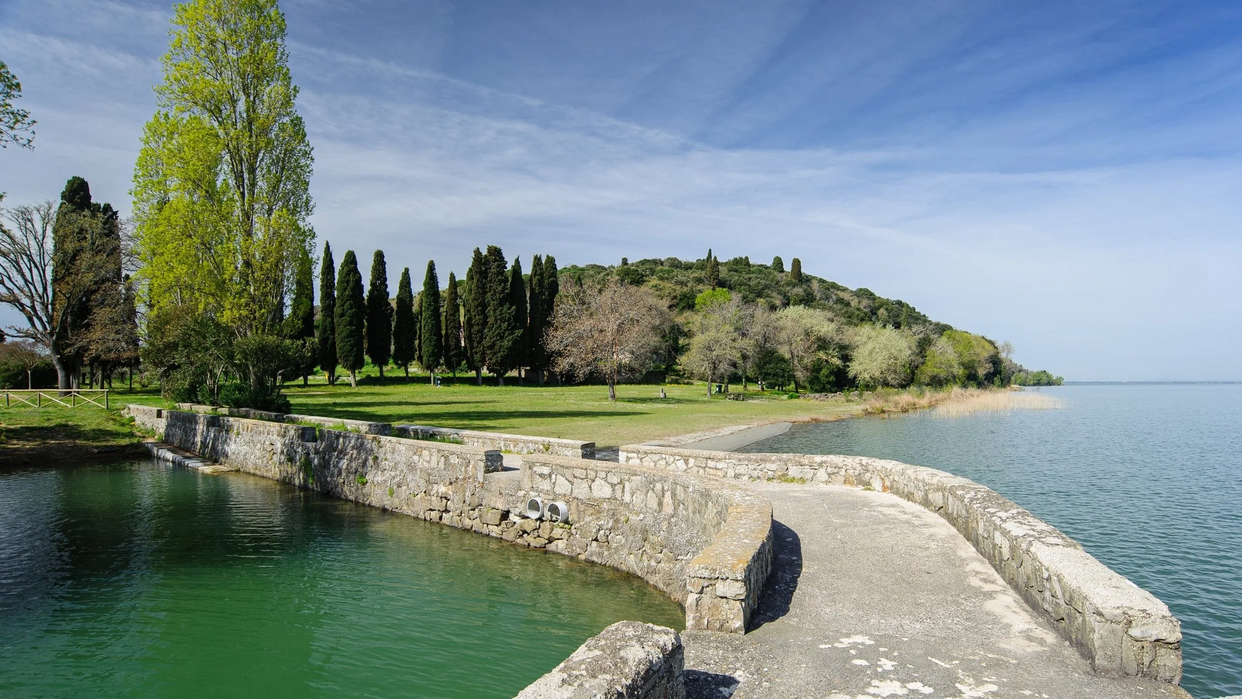 A lakeside park at Isola Polvese with a stone wall and walking path along the water, green trees, and a hill covered with trees in the background under a partly cloudy sky.