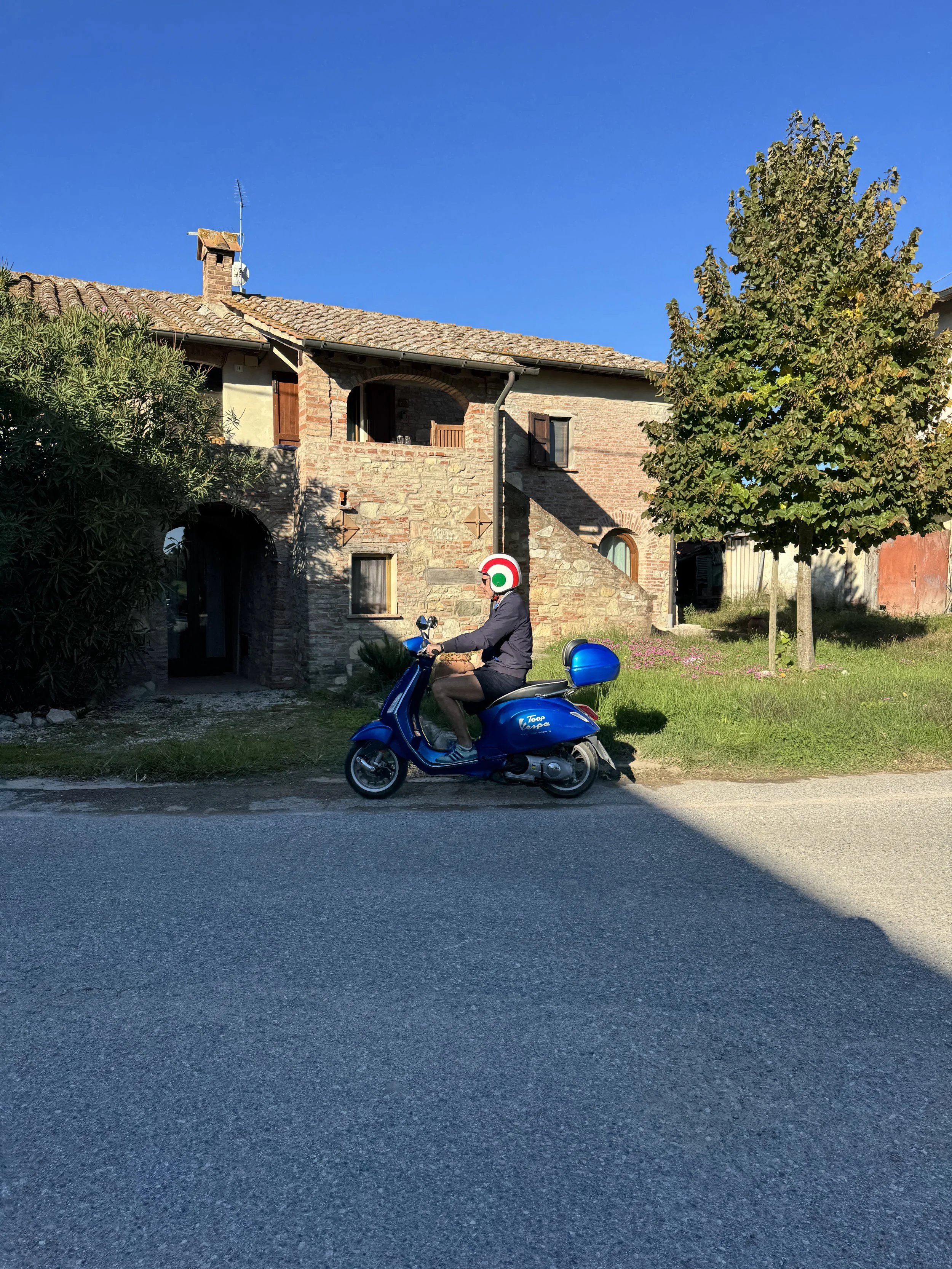 A man riding a blue scooter along a street in front of Casa Castagni, a stone and brick house with a tiled roof. The rider is wearing blue shorts, a blue jacket.  The sky is bright blue.